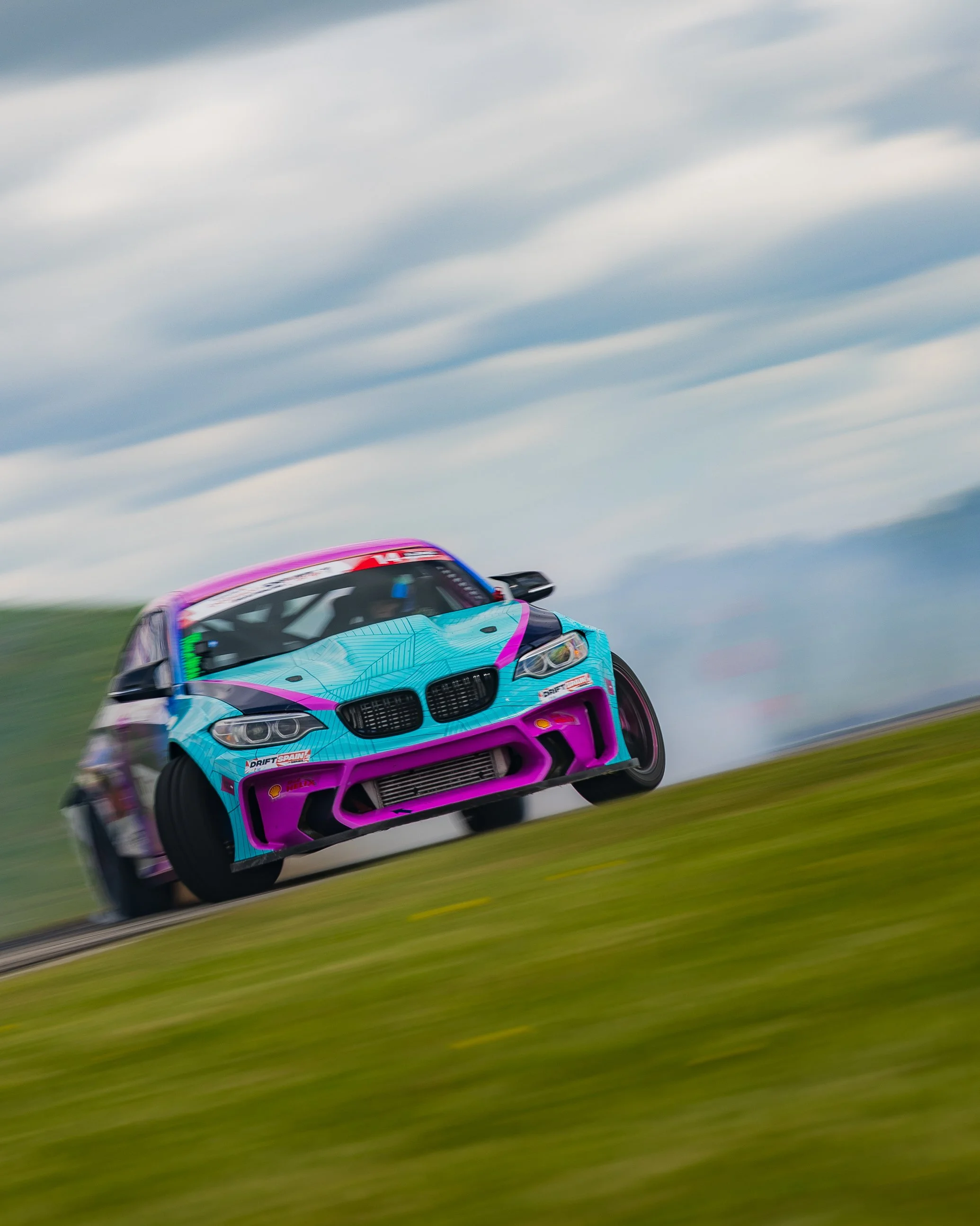 A drift car with a blue, pink, and purple paint scheme racing on a track, kicking up smoke with a cloudy sky in the background.