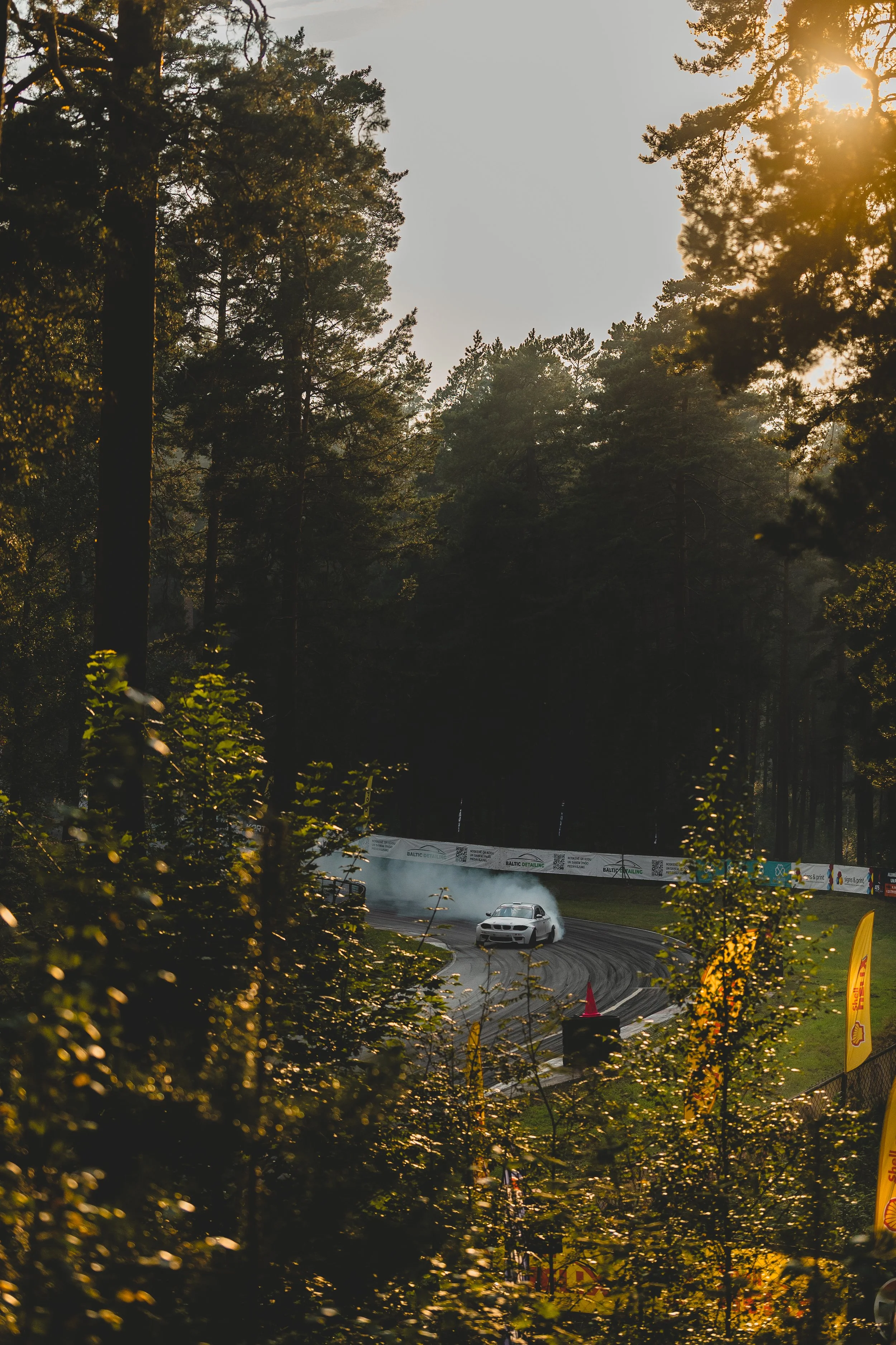 A car drifting on a racetrack surrounded by trees, with smoke and sunlight filtering through the branches.