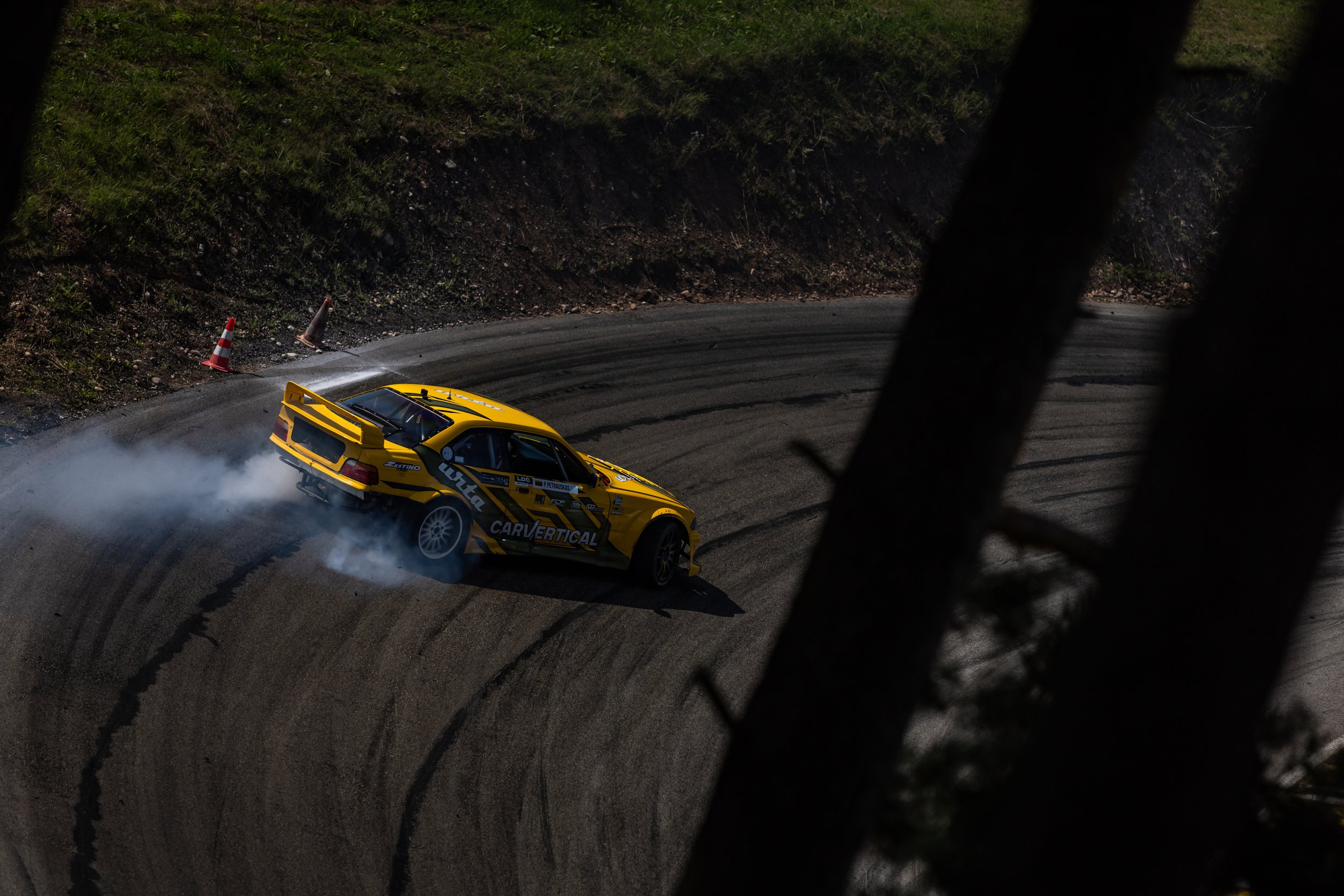 A yellow race car drifts around a bend on a dirt rally stage, creating smoke from its tires, with a grassy embankment and orange cones in the background, partially obscured by a tree trunk in the foreground.