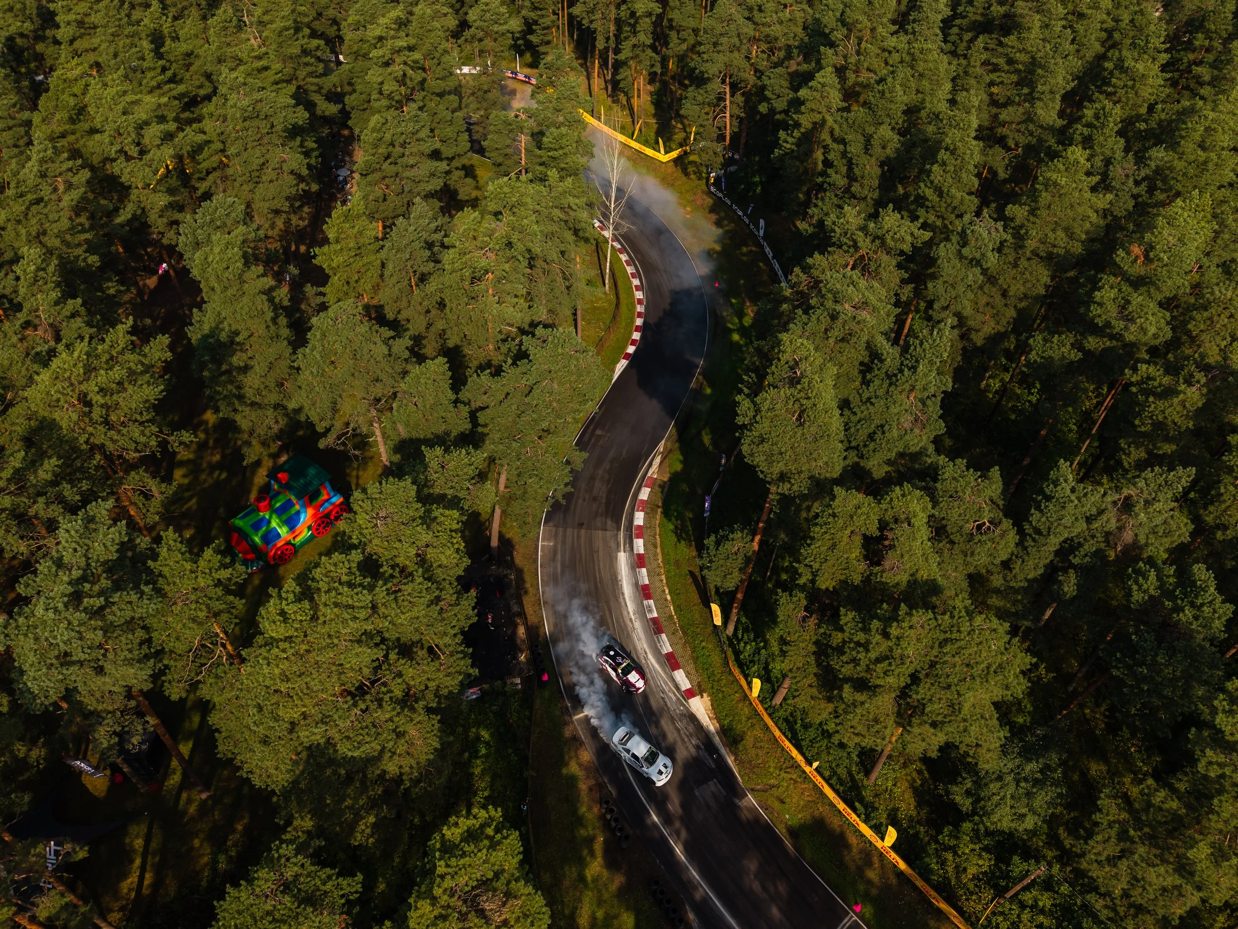 An aerial view of a forested race track with two cars racing and smoke coming from one of them. There is a colorful inflatable train toy visible among the trees on the left side.
