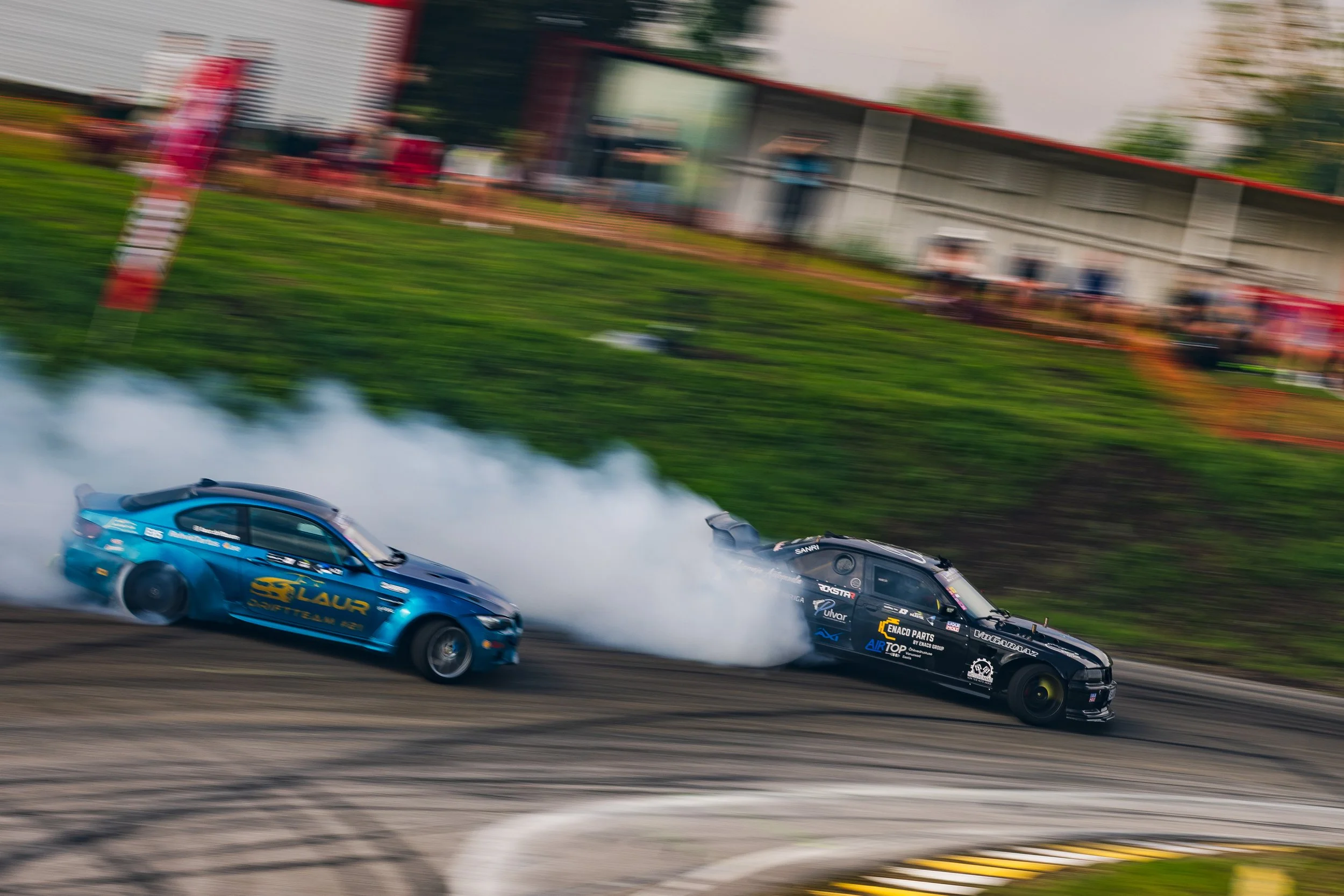 Two race cars drifting on a track, with smoke rising from their tires as they navigate a turn, blurred spectators in the background.