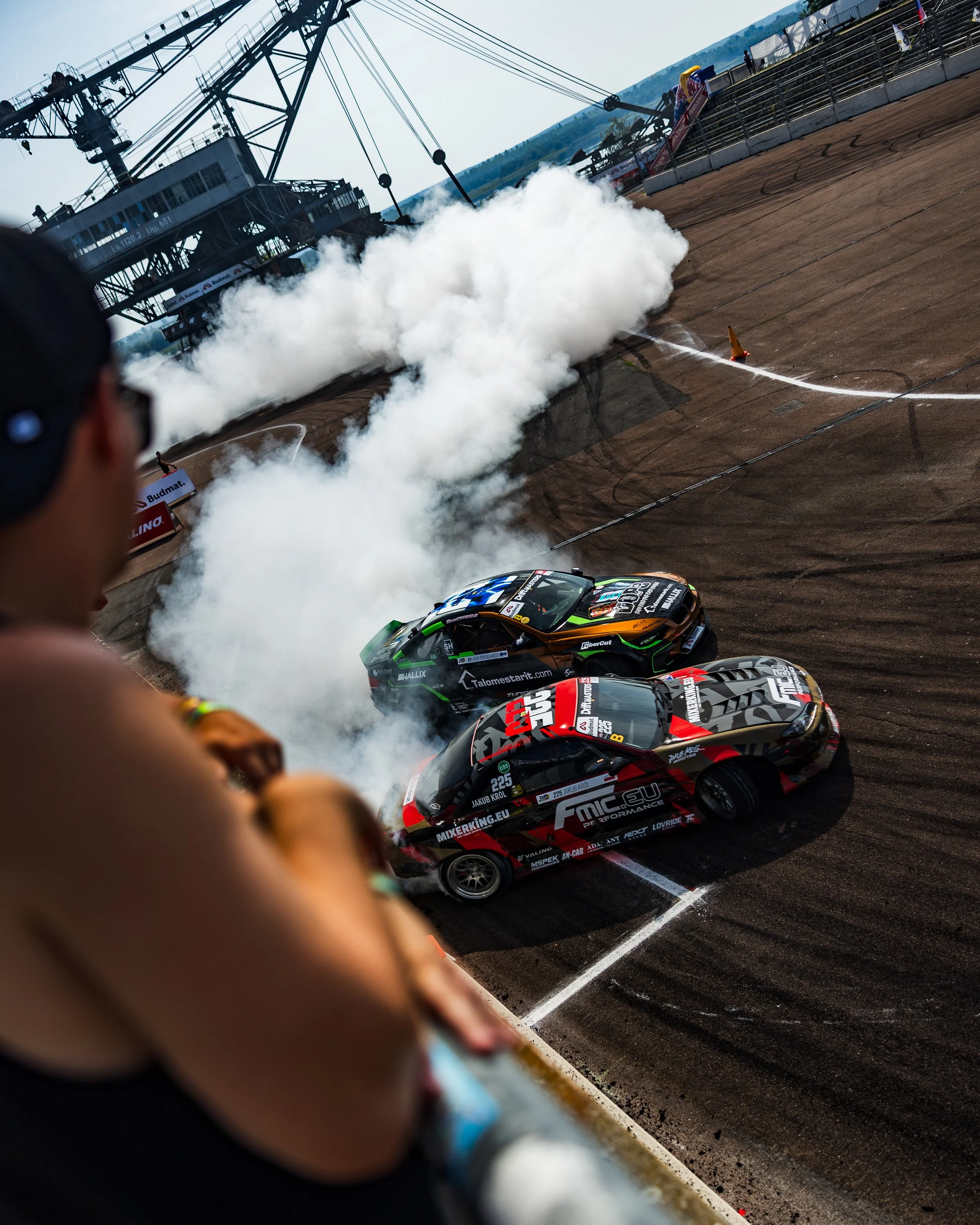 Two race cars spinning out and creating smoke on a dirt racing track during a race, with a spectator leaning over the railing in the foreground.
