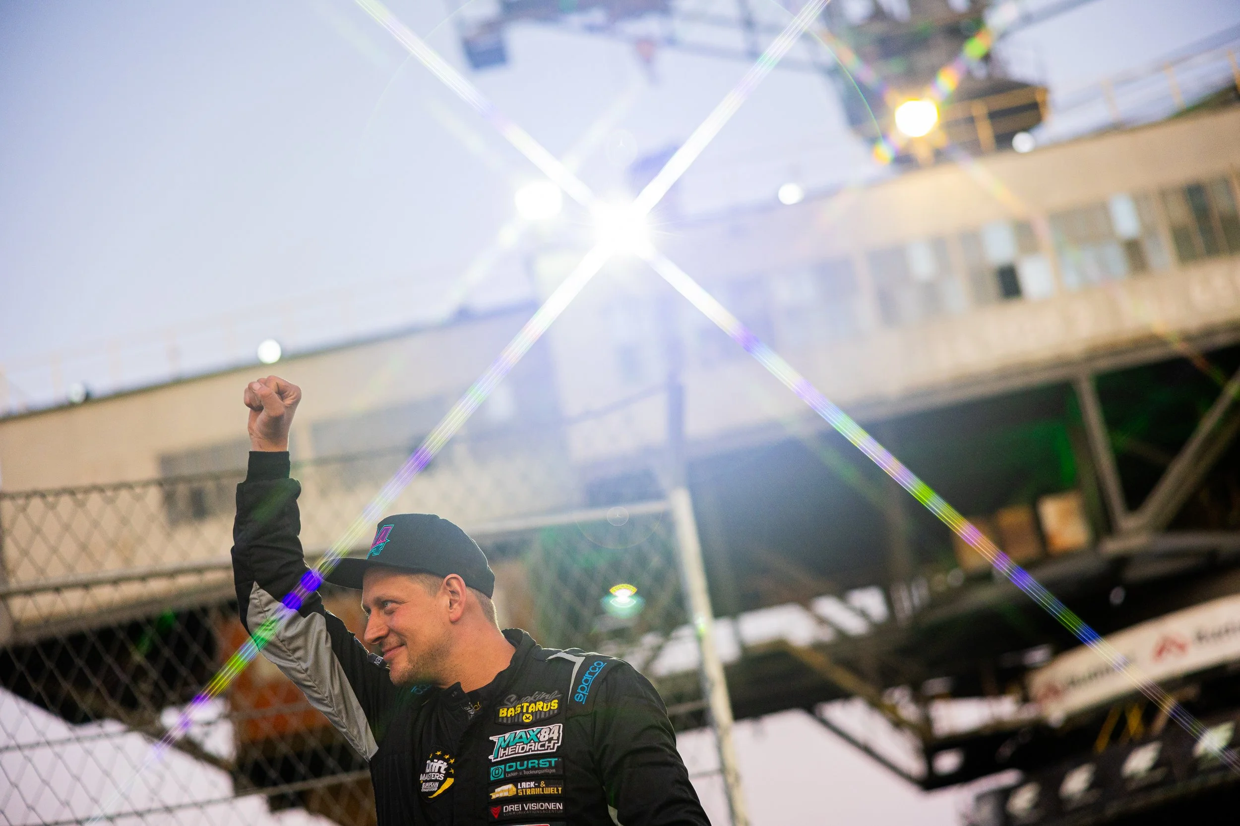 A race car driver in a black racing suit and cap smiling and raising his fist in victory, with sunlight creating starburst effects in the background.