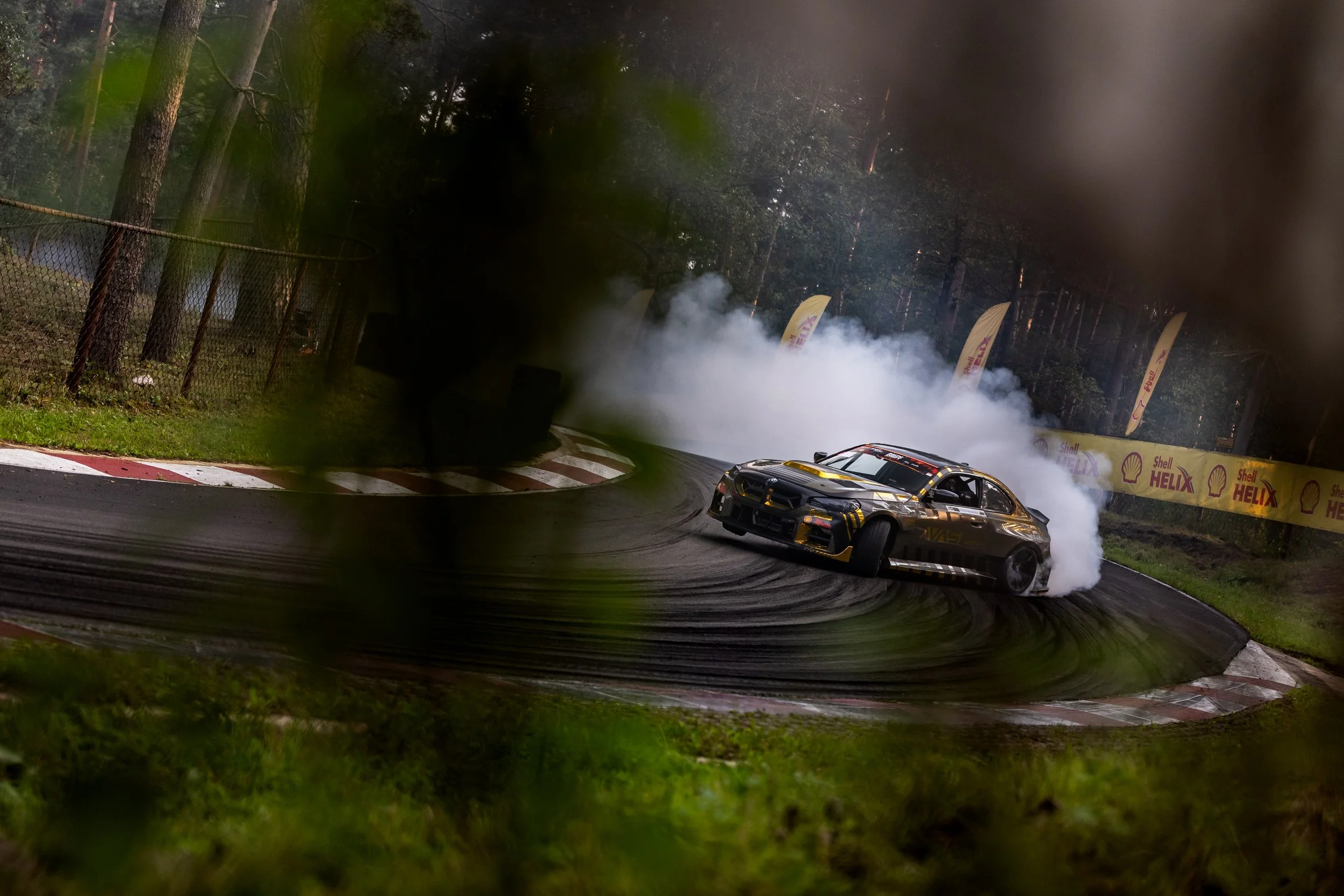 A racing car performs a drift on a racetrack, creating a large cloud of smoke behind it, with trees and banners in the background.