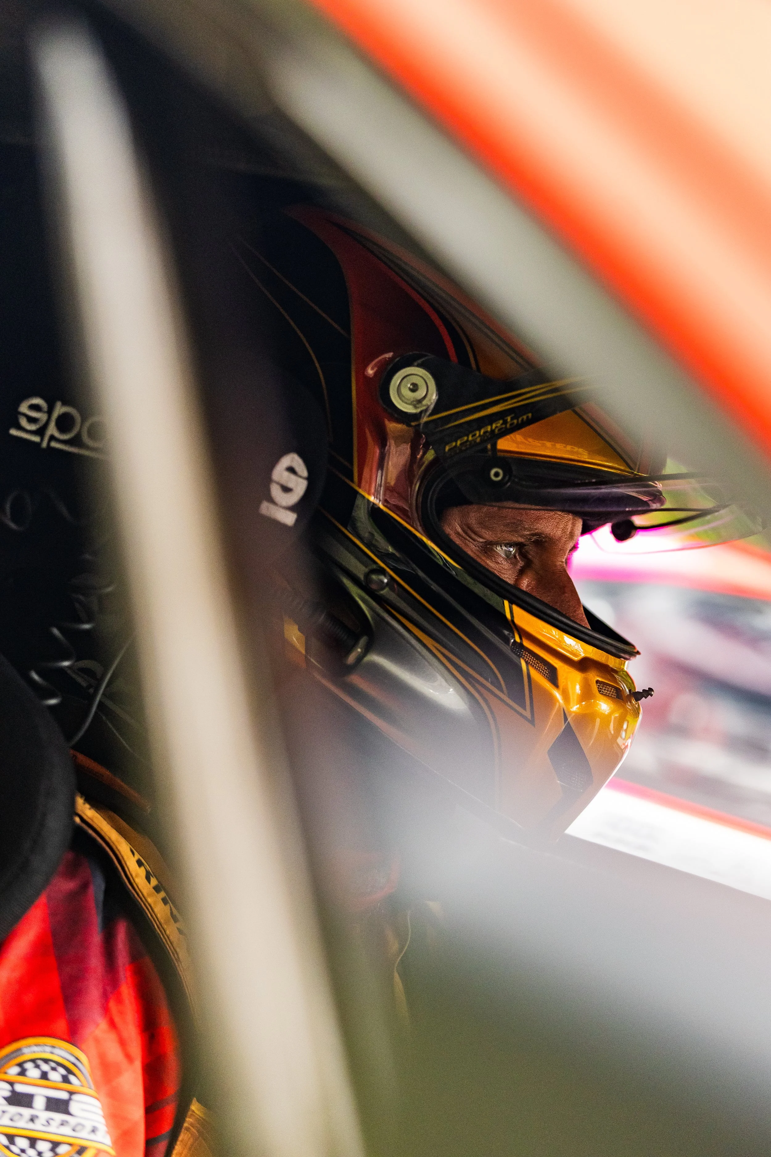 Close-up of a race car driver wearing a yellow helmet and racing suit, sitting inside a car, seen from the side through an opening in the vehicle.