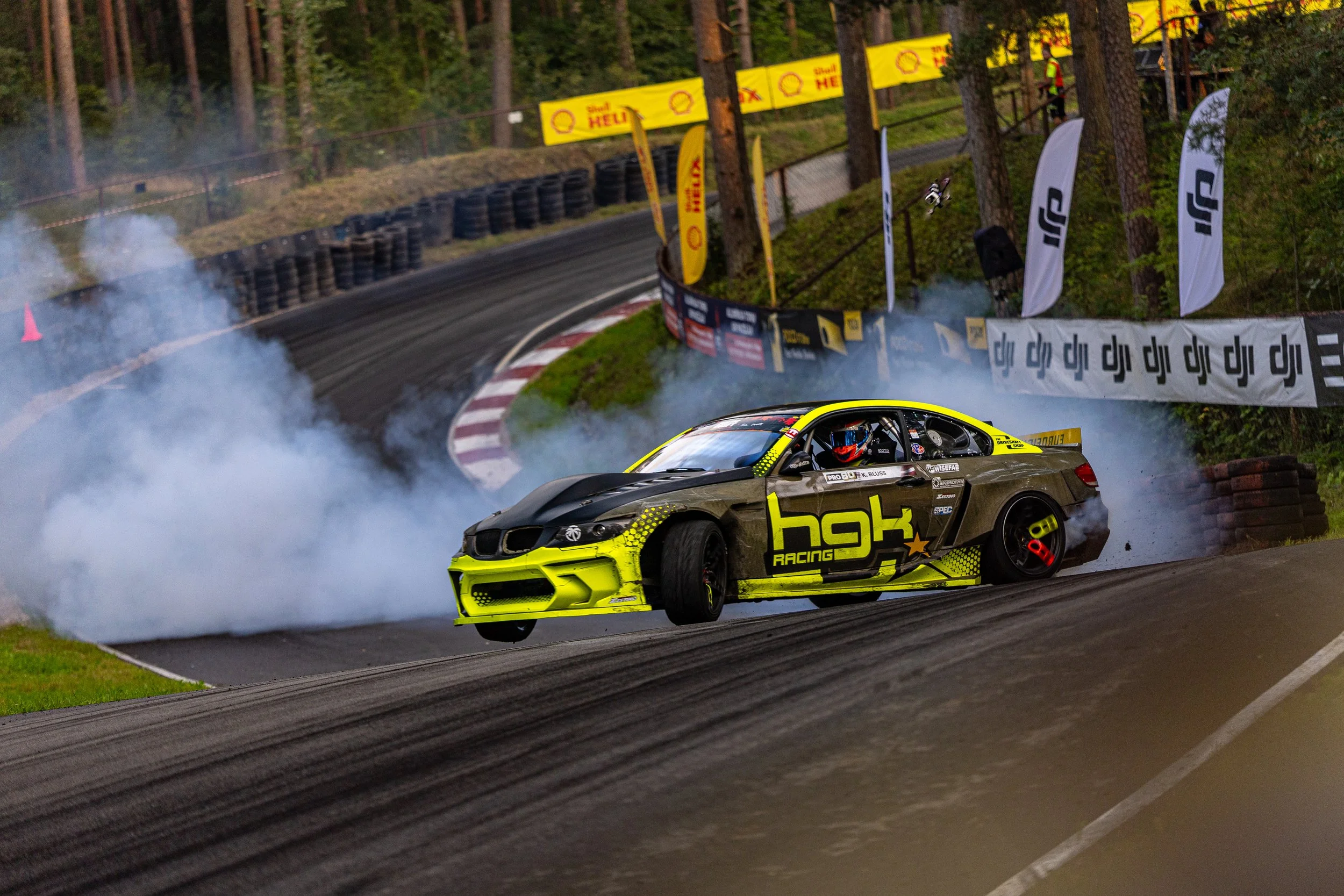 A race car drifting on a racetrack, creating smoke with skid marks, surrounded by banners and trees.