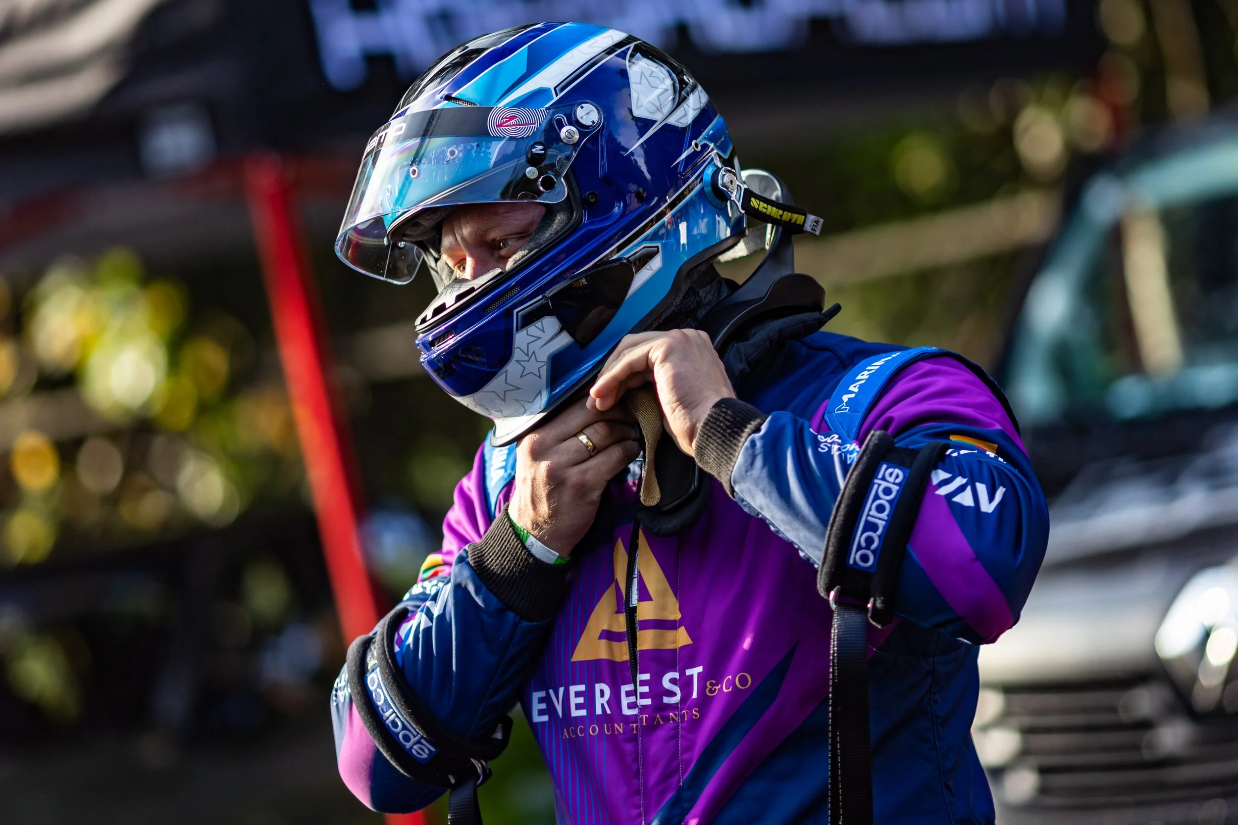 A race car driver wearing a blue and purple racing suit and a blue helmet, adjusting his collar outdoors.