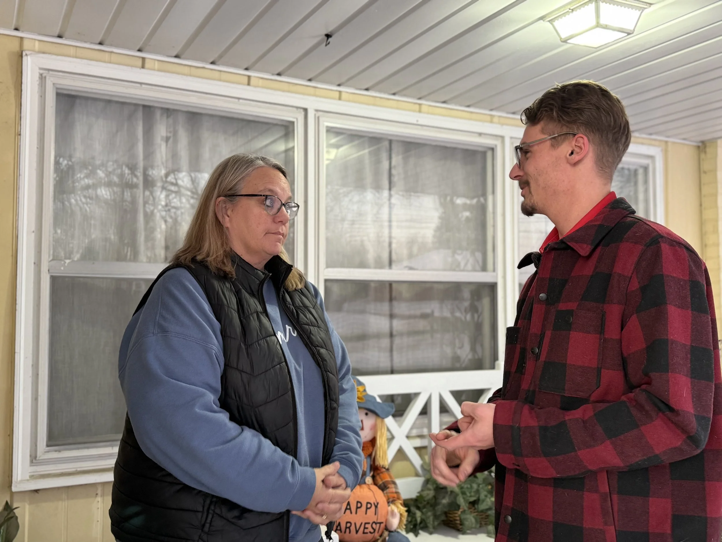 A woman and a man are standing face-to-face on a porch, engaging in conversation. The woman has shoulder-length hair, glasses, and is wearing a blue sweatshirt with a black vest. The man has short hair, glasses, and is dressed in a red and black checkered shirt. There is a decorative scarecrow and some potted plants on a table behind them.