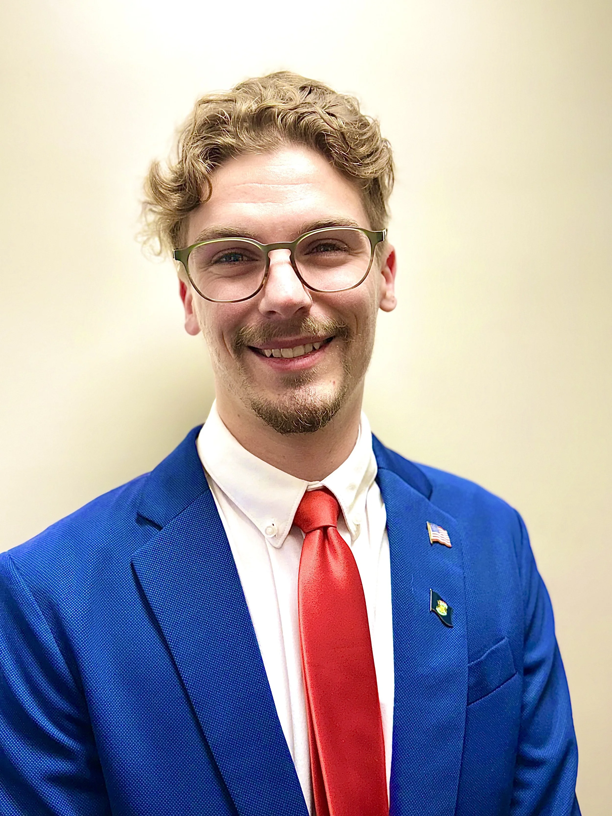 A smiling man with curly blond hair, glasses, and facial hair, wearing a blue suit, white shirt, and red tie, with American and other patriotic pins, standing against a plain beige background.