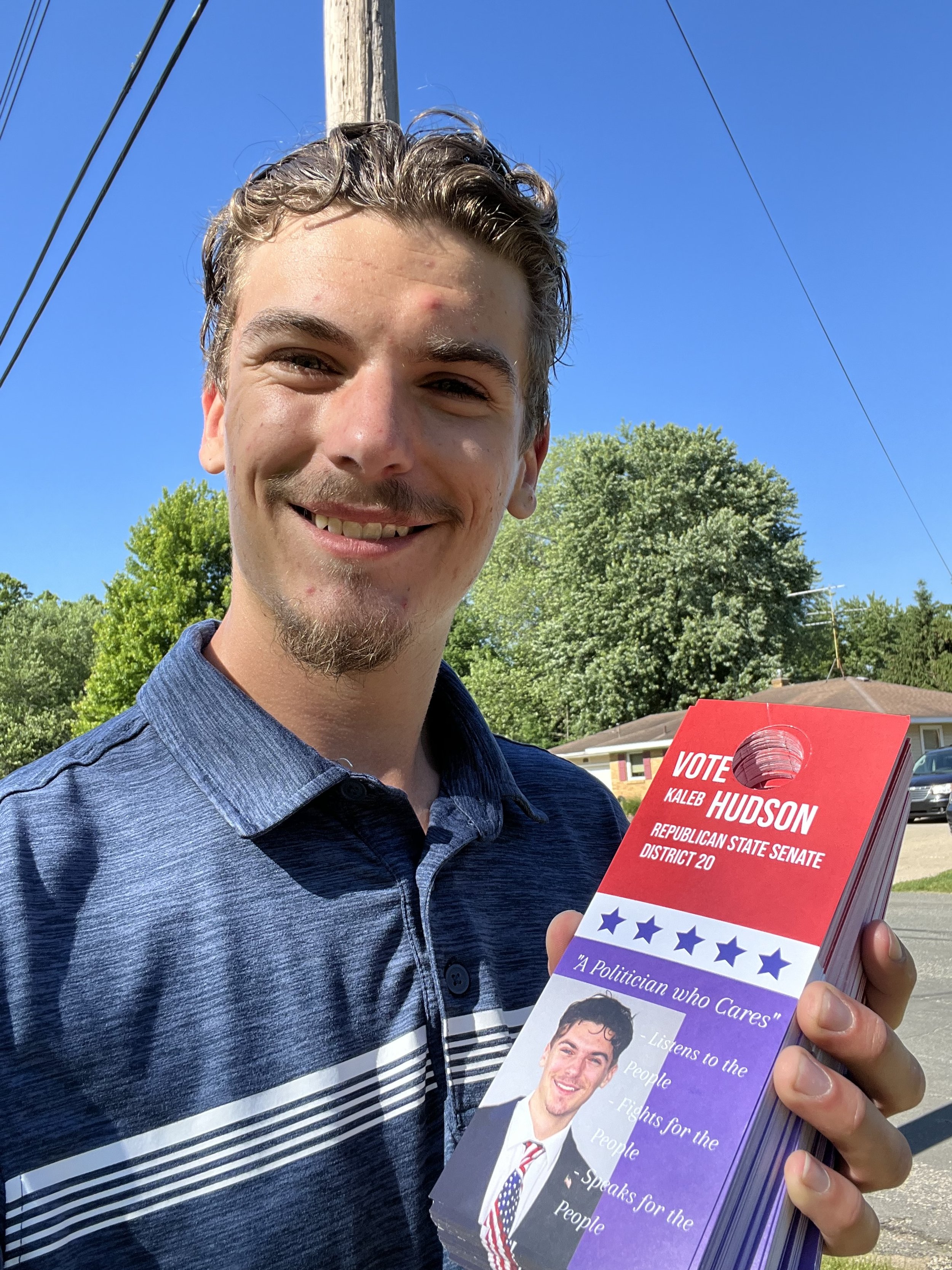A young man smiling outdoors, holding campaign flyers for Kaleb Hudson, Republican State Senate candidate.