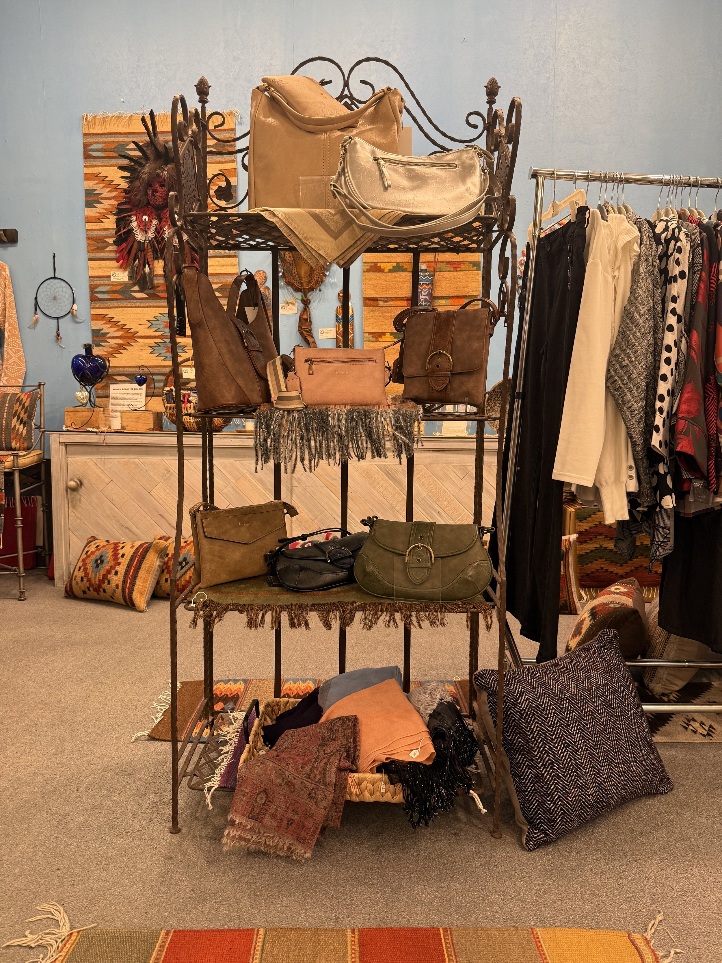 Display of handbags, clothing, scarves, and textiles in a boutique shop with woven wall art and decorative pillows.