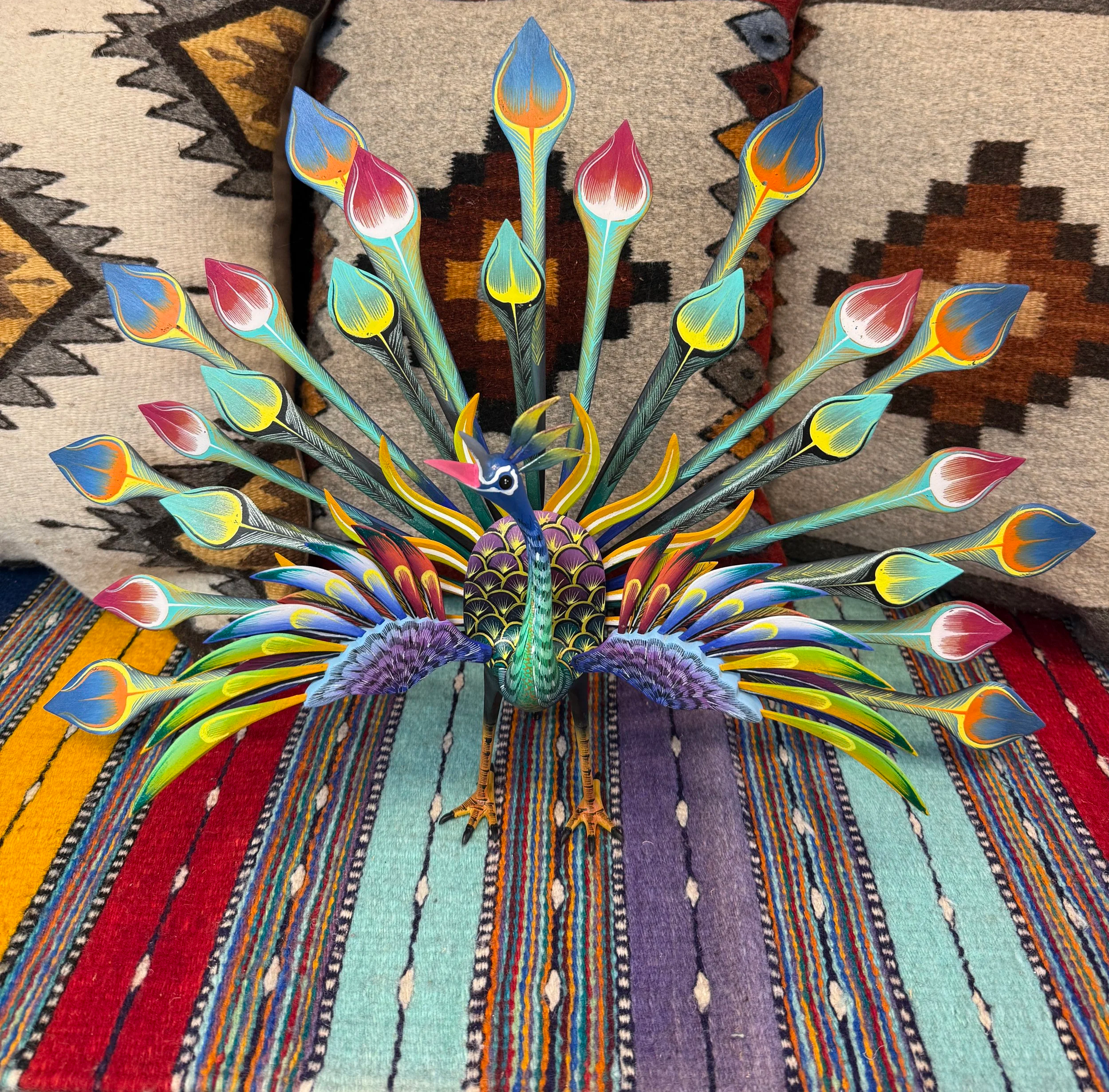 Colorful handmade wooden carving of a peacock with spread feathers, on a multicolored woven textile background.