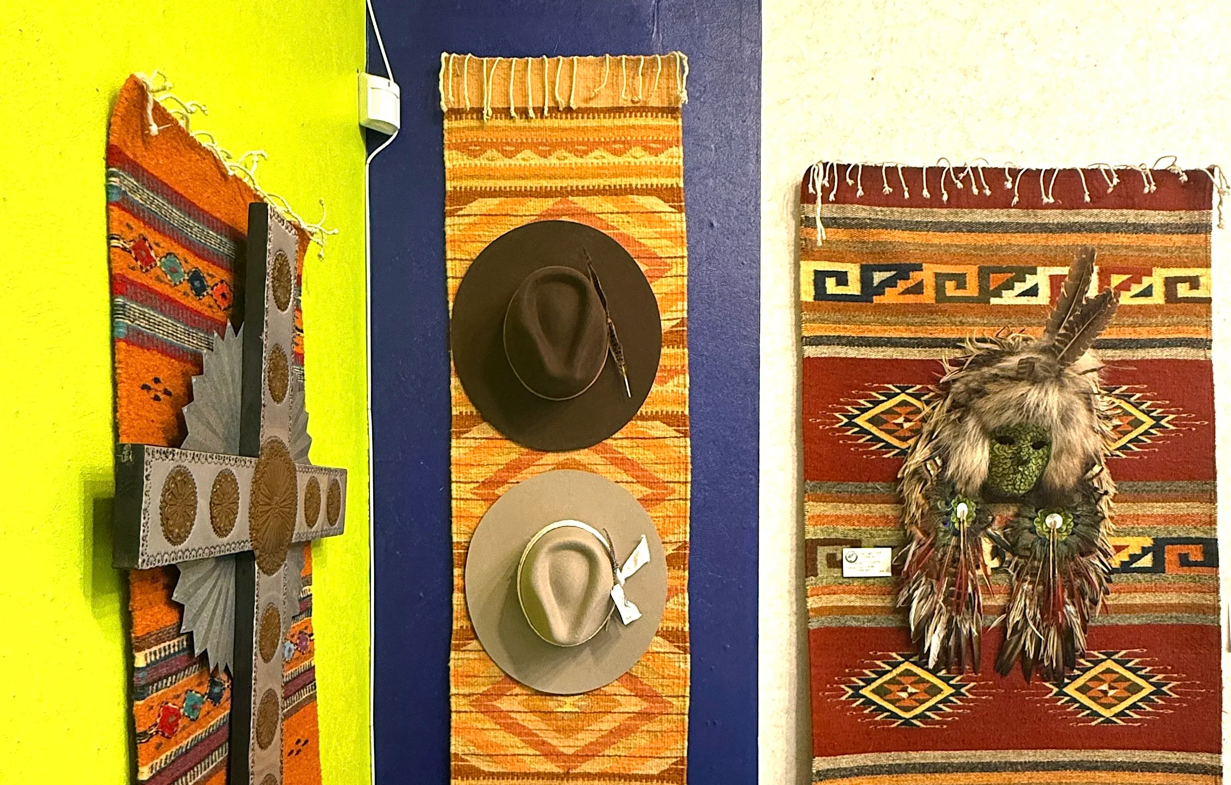 Display of hats on a colorful striped wall with embroidered rugs and Native-American decorative items, including a feathered mask and clothing rack with various shirts, jackets, sweaters, and cardigans.