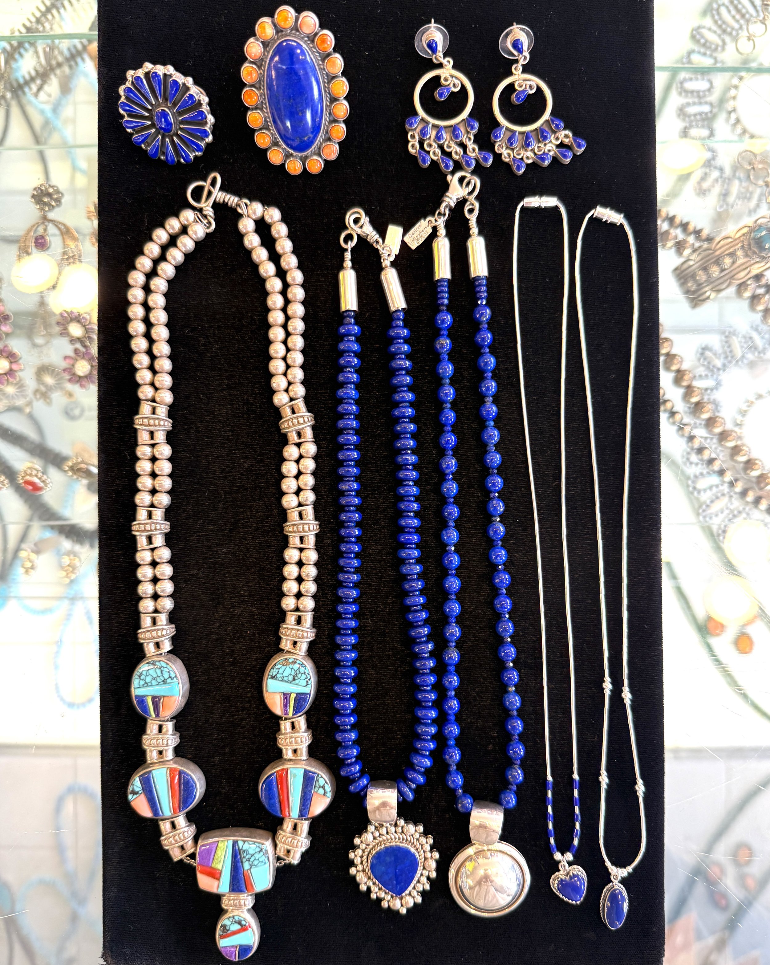 Collection of lapis lazuli jewelry including necklaces, earrings, and chains with blue, turquoise, and silver accents displayed on a black background.