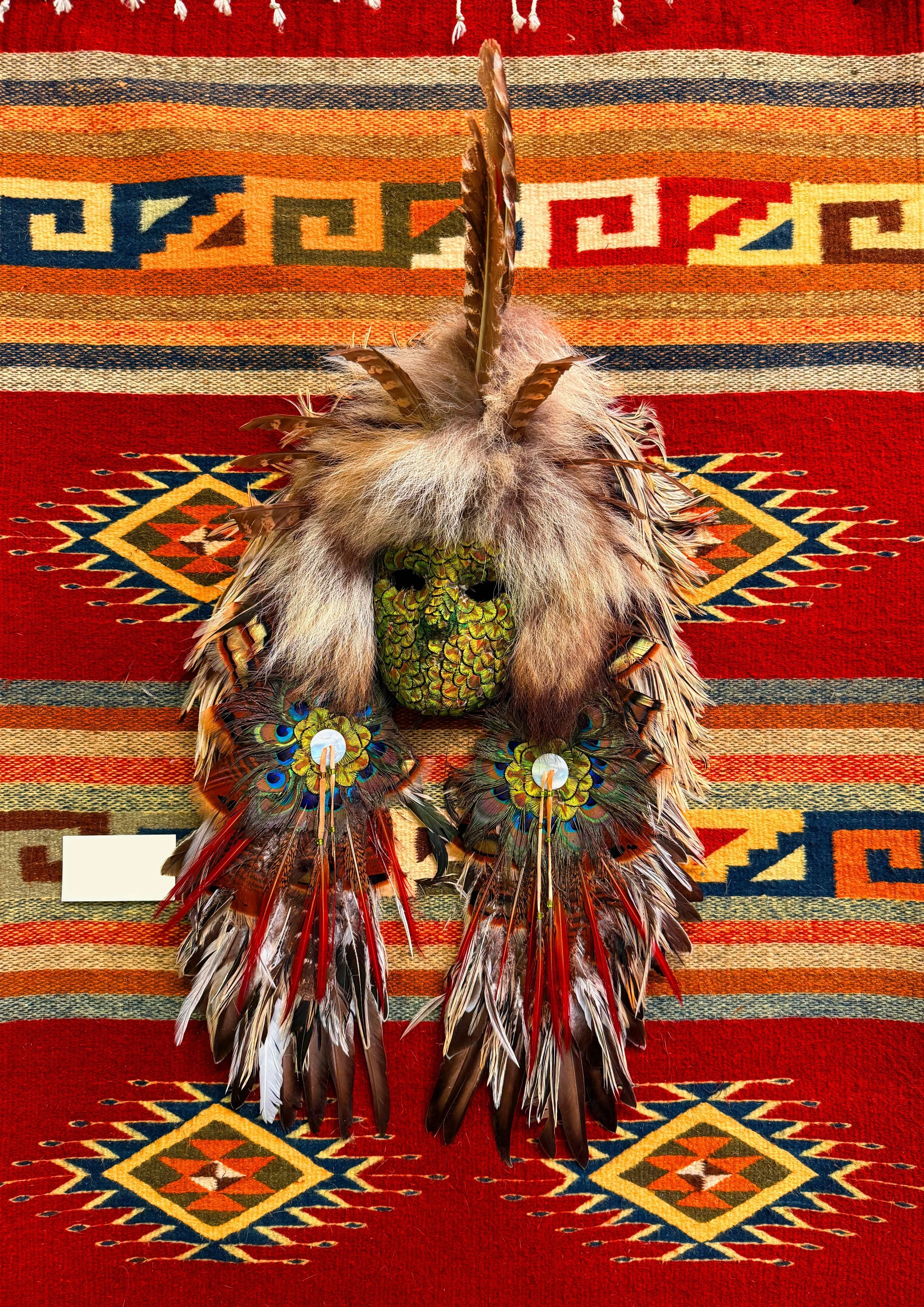 An authentic handmade Native-American mask in many colors with a handwoven rug behind it.