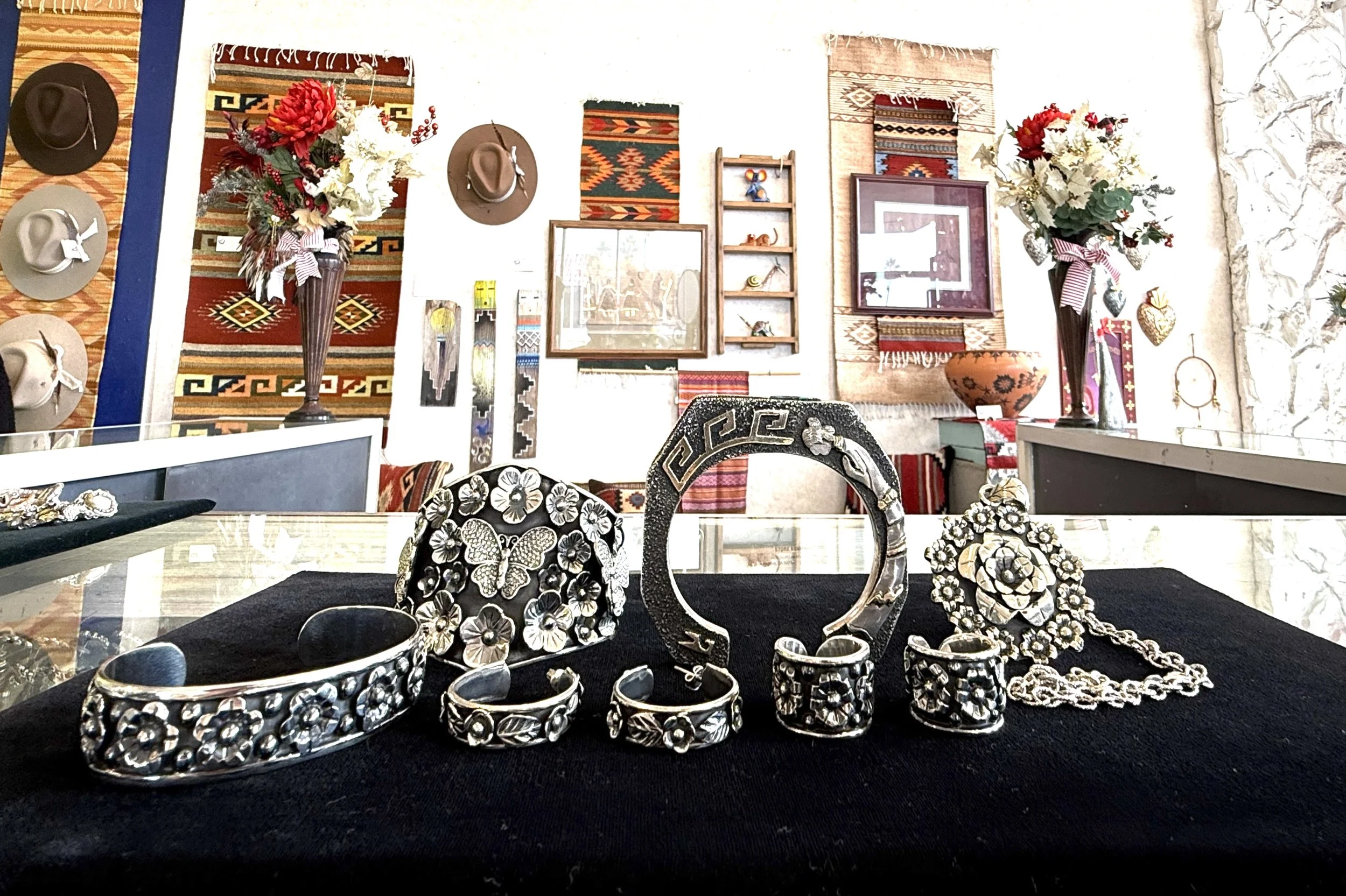 Silver jewelry including bracelets, rings, and earrings with floral and butterfly designs displayed on a black fabric surface, with a decorated room featuring rugs, hats, and floral vases in the background.