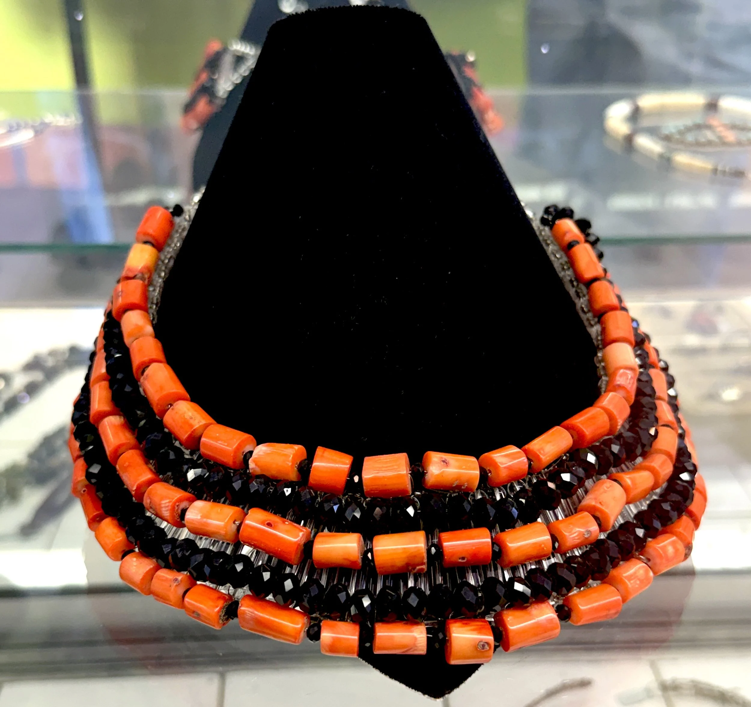 Beaded necklace with orange rectangular beads and black faceted beads displayed on a black stand.