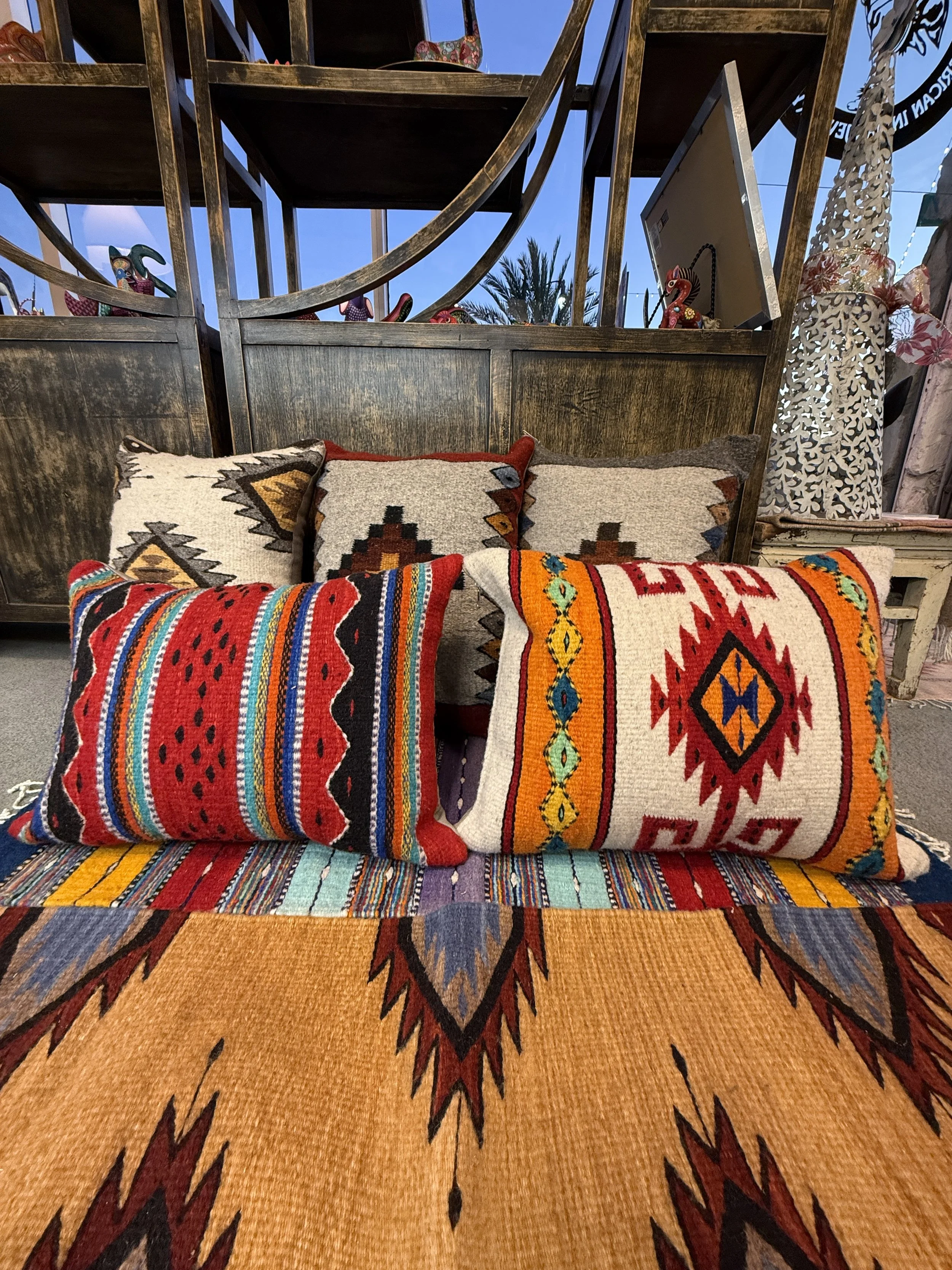 A home décor display of decorative throw pillows with vibrant patterns, arranged on a bed or lounge area with a woven textile, against a background of wooden shelves with pottery and decorative items.