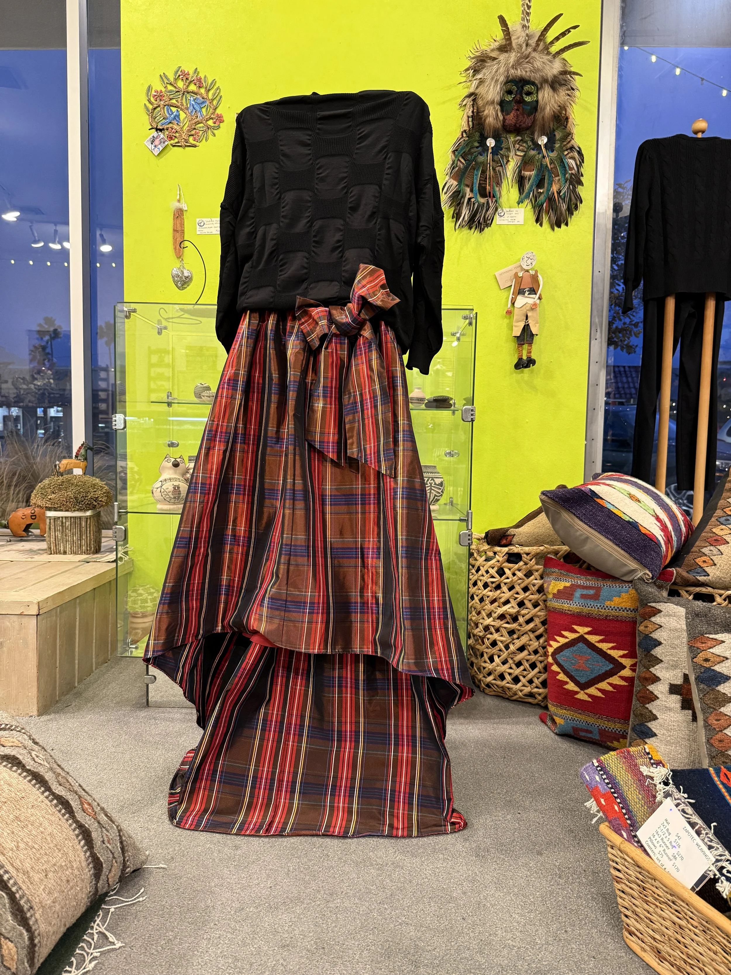 A mannequin dressed in a black top and a long plaid skirt, with a large bow in the front, displayed with colorful wall decor and textiles around.