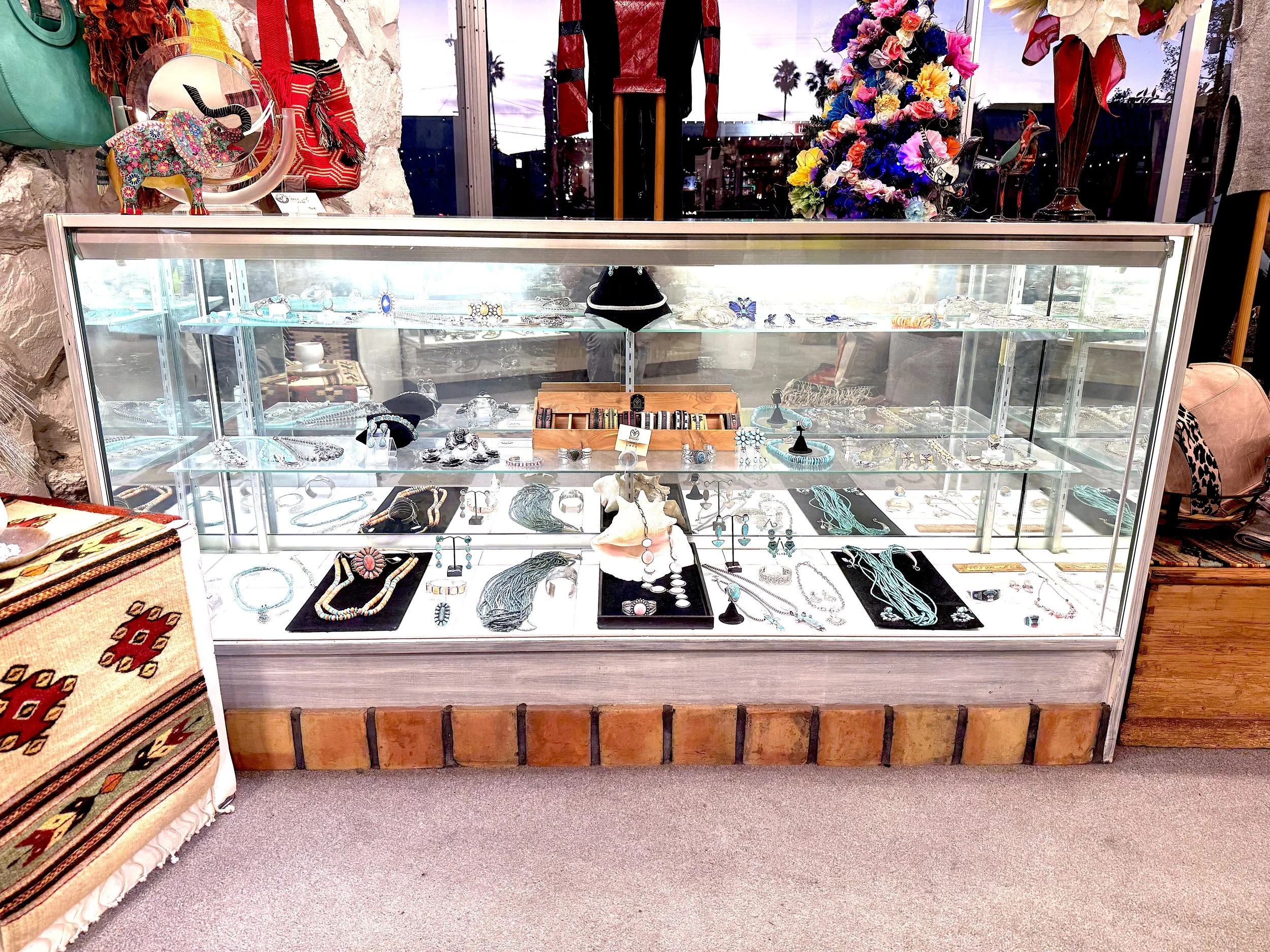 Jewelry display case filled with various necklaces, bracelets, earrings, and rings, with decorative items and accessories around it. A colorful floral arrangement and decorative elephant sculpture are on top of the display case.