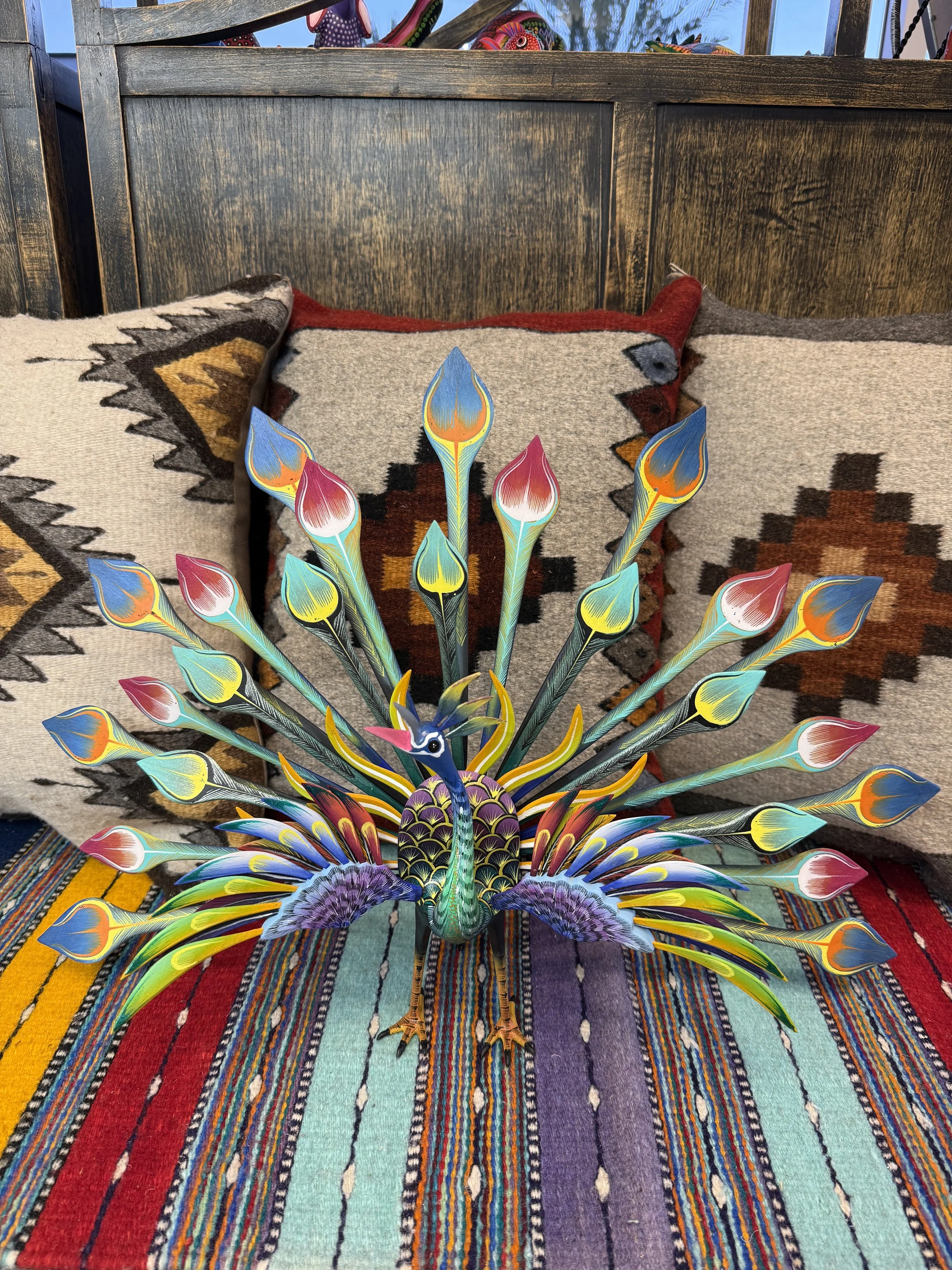 Colorful decorative folk-art peacock with multiple tails, placed on a striped fabric surface with patterned cushions in the background.