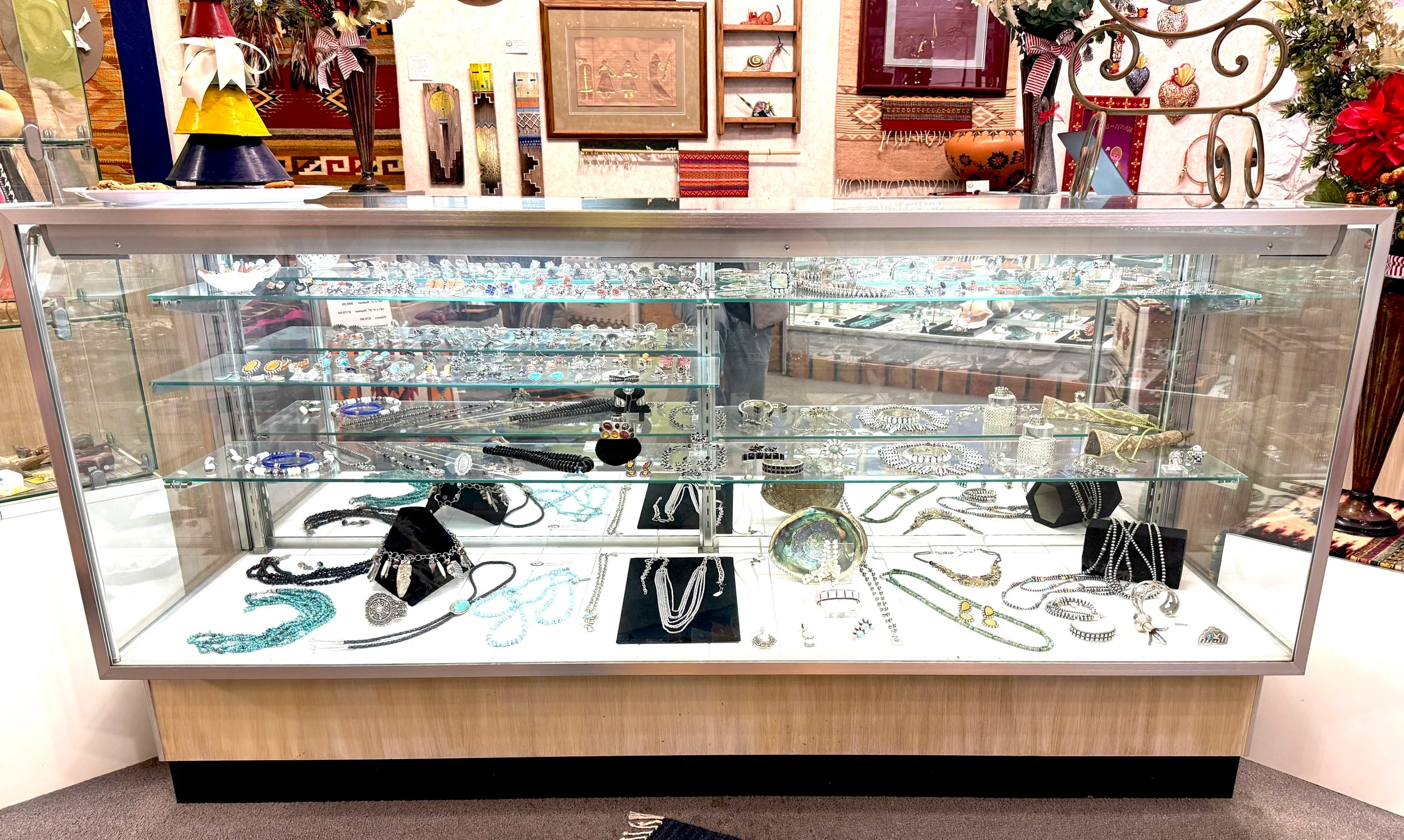 Jewelry display case with various necklaces, bracelets, and rings inside, located in a shop with decorative artwork and items in the background.