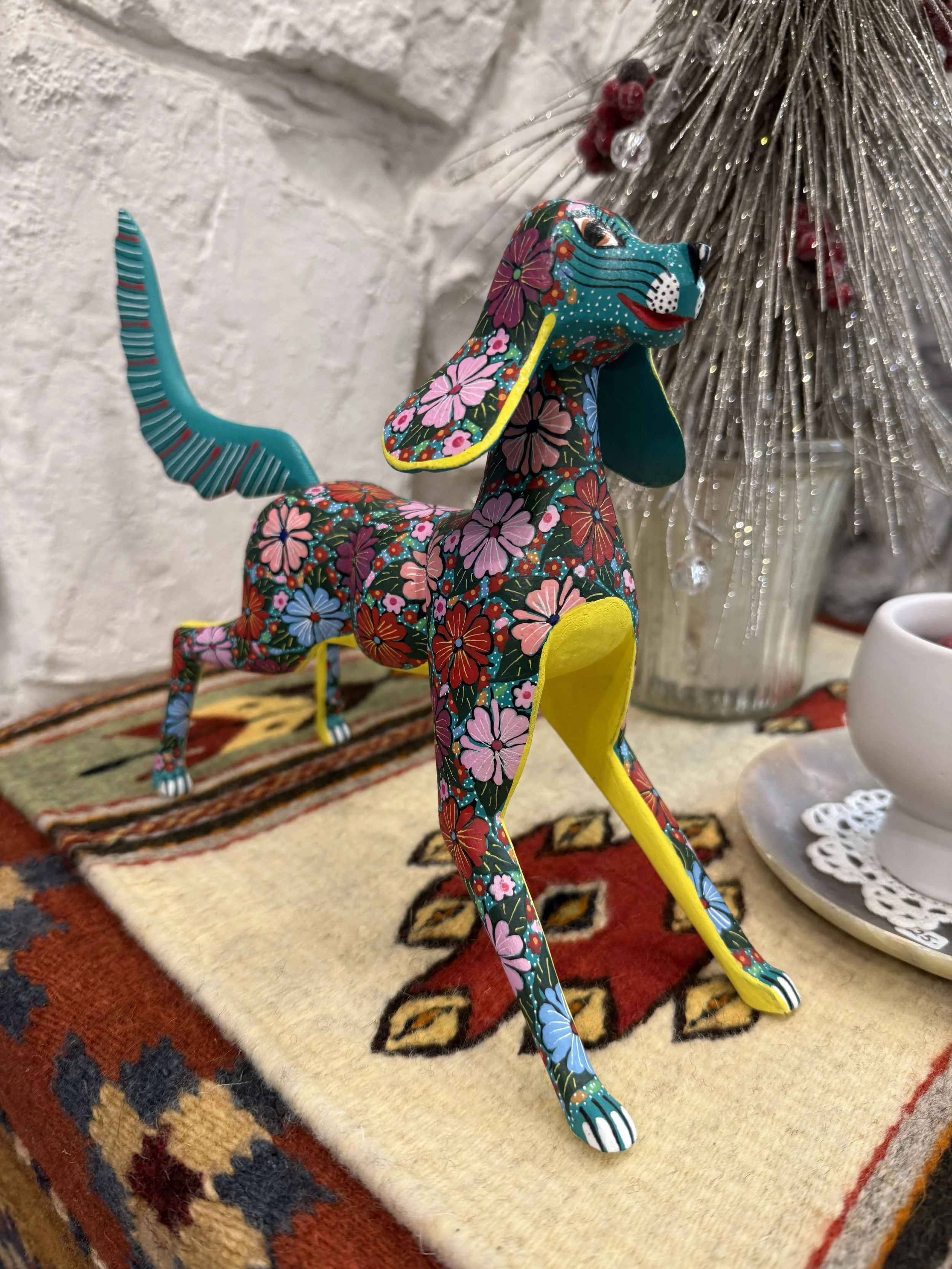 Colorful decorative dog figure with floral pattern, yellow nose, blue ears, and striped tail, standing on a patterned rug near a white ceramic cup and a silver holiday decoration.