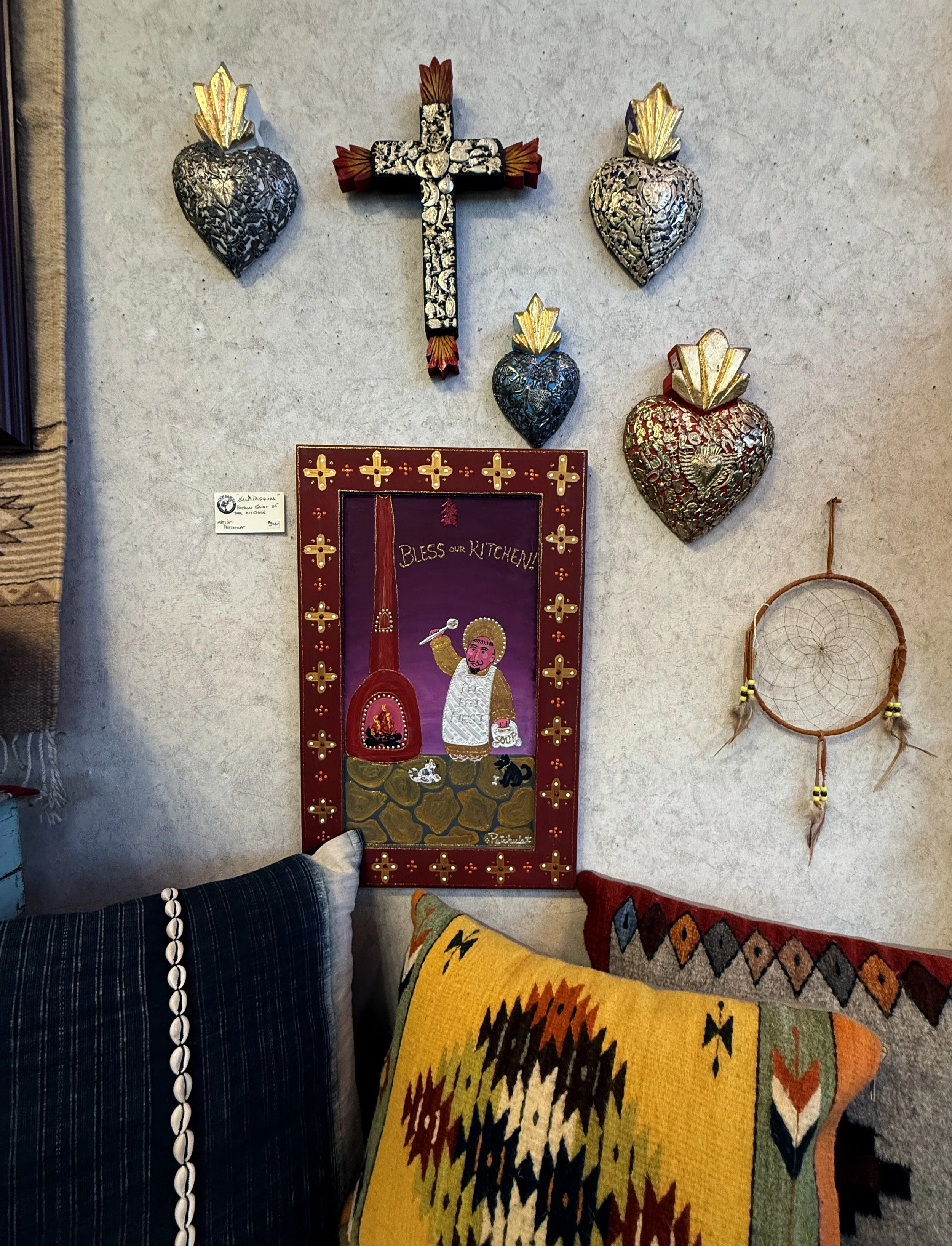 Decorative wall with a cross, four metallic heart-shaped ornaments, a framed painting, and a dreamcatcher. Part of a couch with colorful patterned pillows is visible at the bottom.