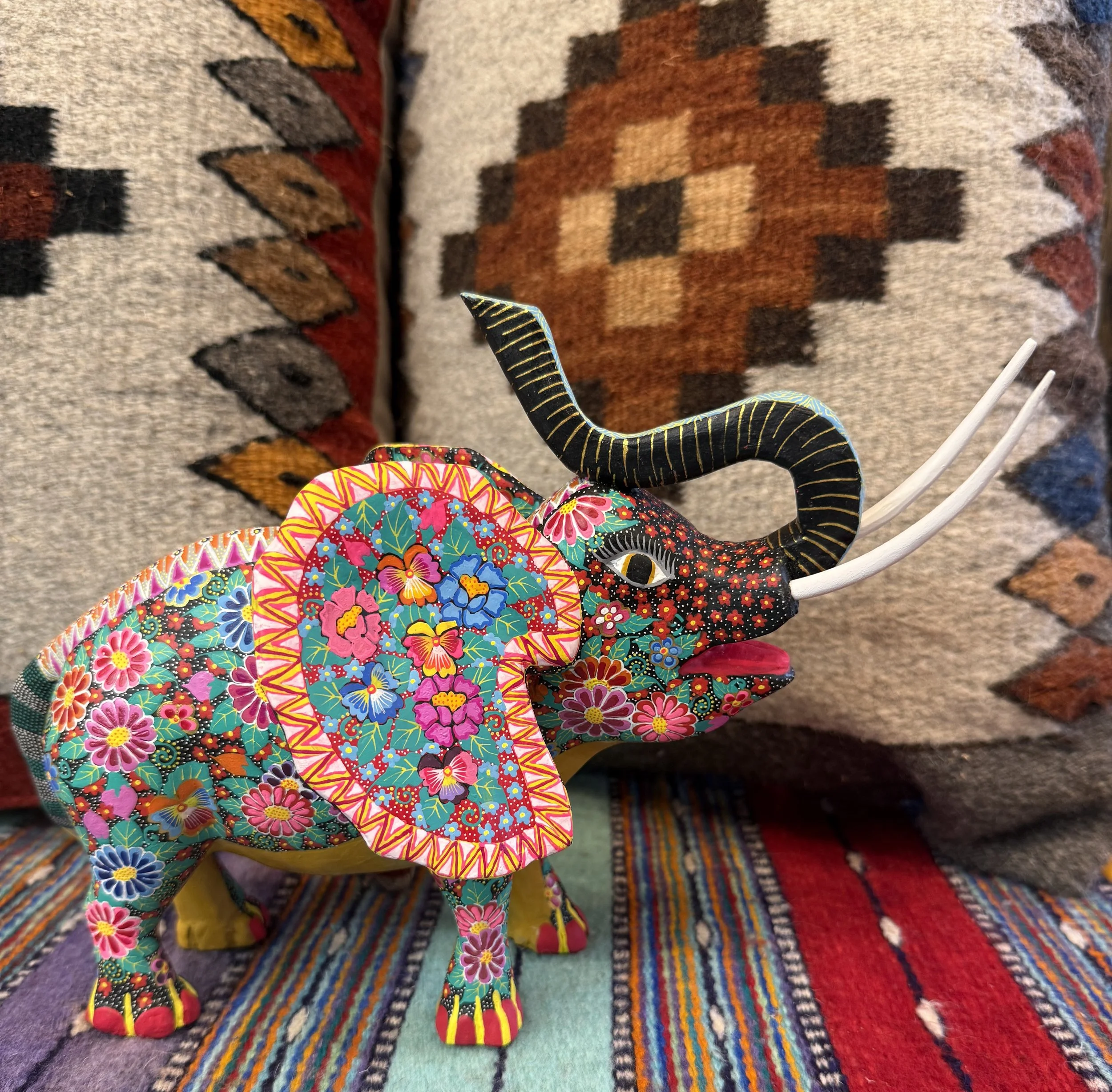 Colorful handmade decorative elephant figurine with floral patterns, black and yellow striped trunk, and white tusks, placed on a multicolored striped textile with a patterned woven textile in the background.