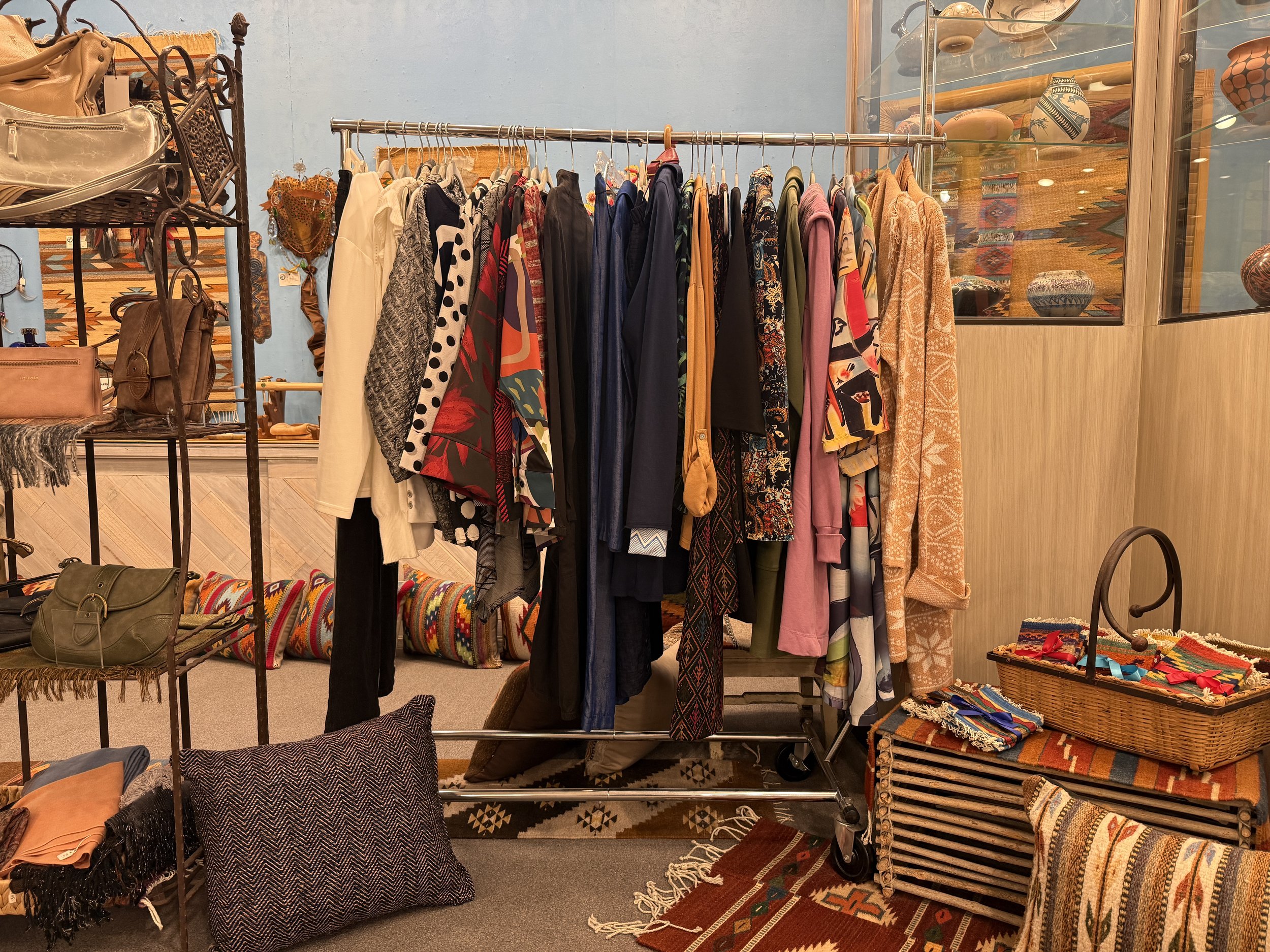 Clothing rack with colorful dresses, tops, and jackets, surrounded by woven baskets and textile decor in a boutique.