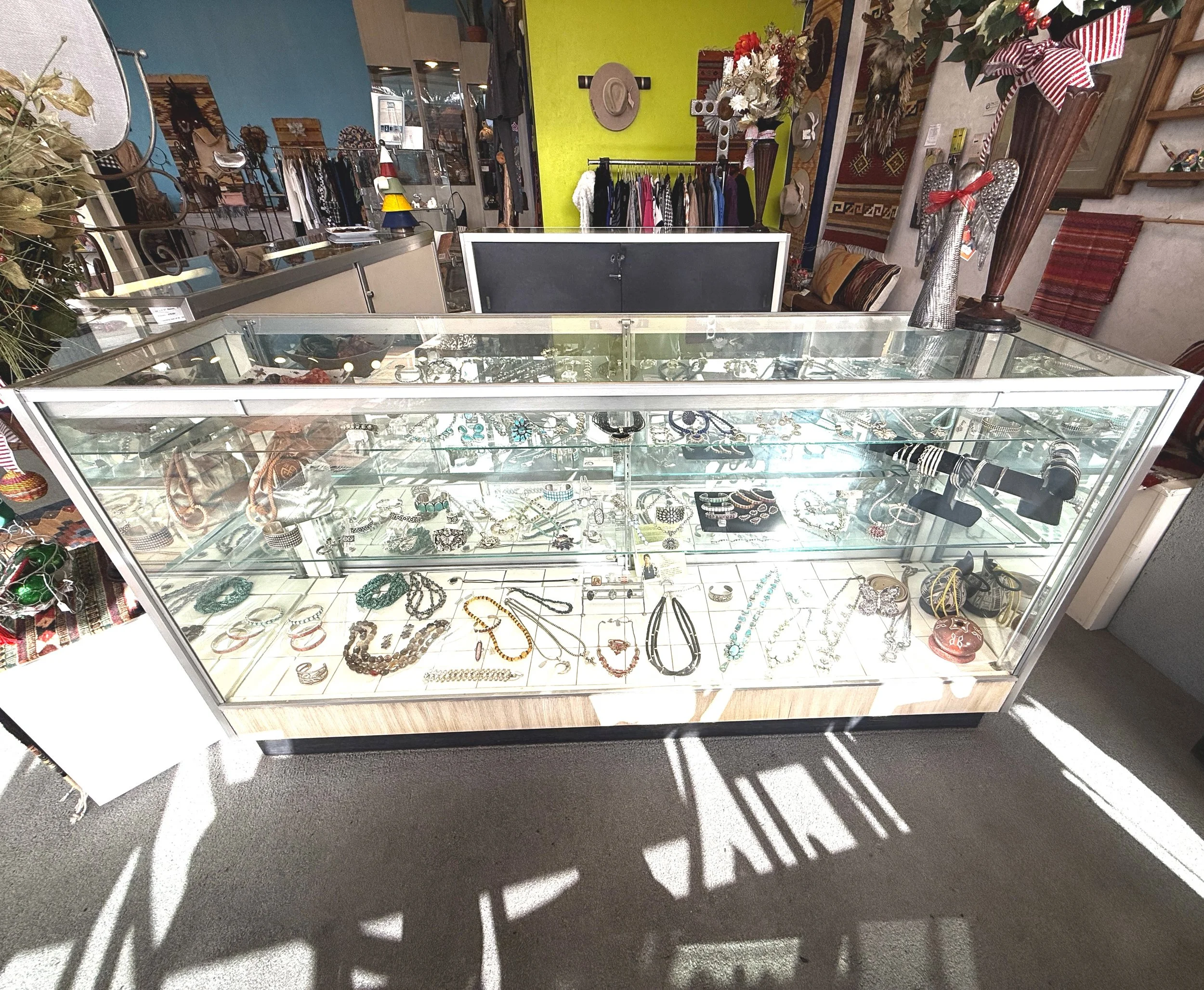 A glass display case filled with various jewelry, including necklaces, bracelets, rings, and earrings, in a store, Moon Dancer. Behind the case, there are clothing racks, colorful walls, and decorative items such as flowers and holiday ornaments.