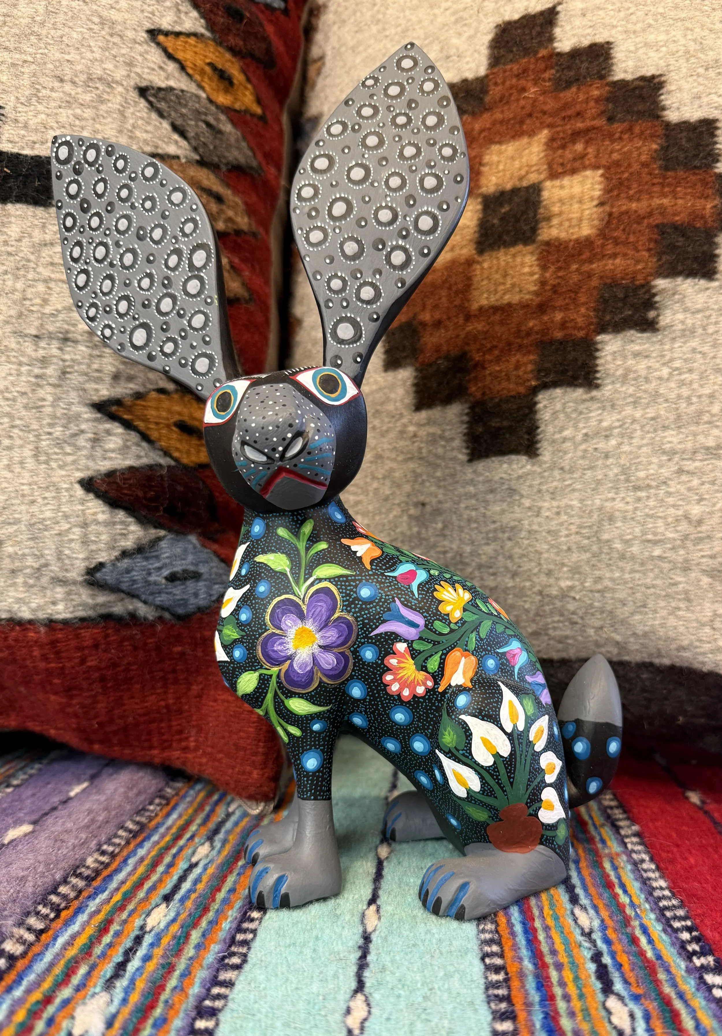 Colorful decorative rabbit figurine with large, patterned ears, sitting on a multicolored striped fabric, against a woven pillow background.