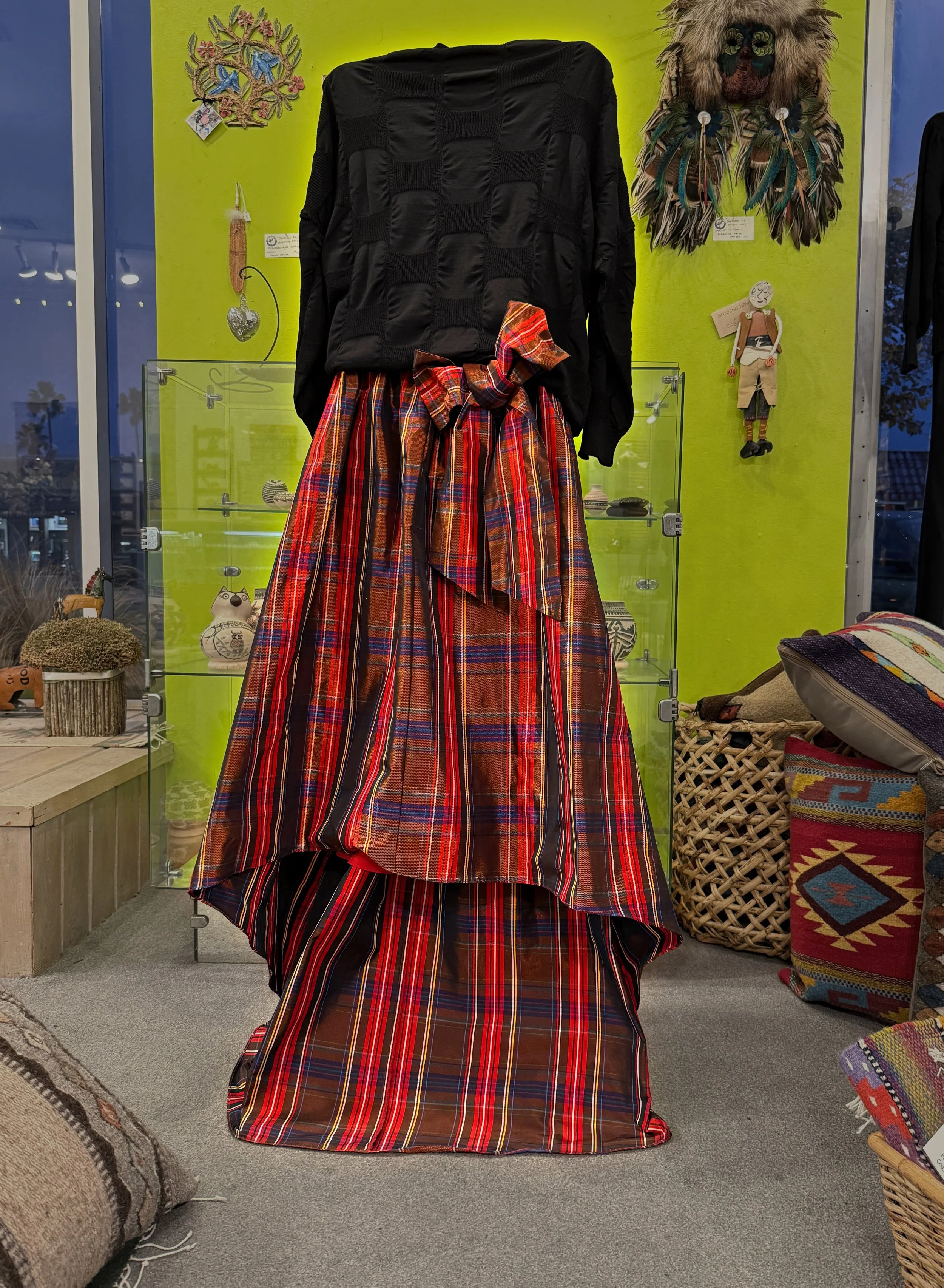 Display of a black knit top and a long red plaid skirt in Moon Dancer in Redondo Beach. In the background are decorative wall hangings and other handcrafted items.