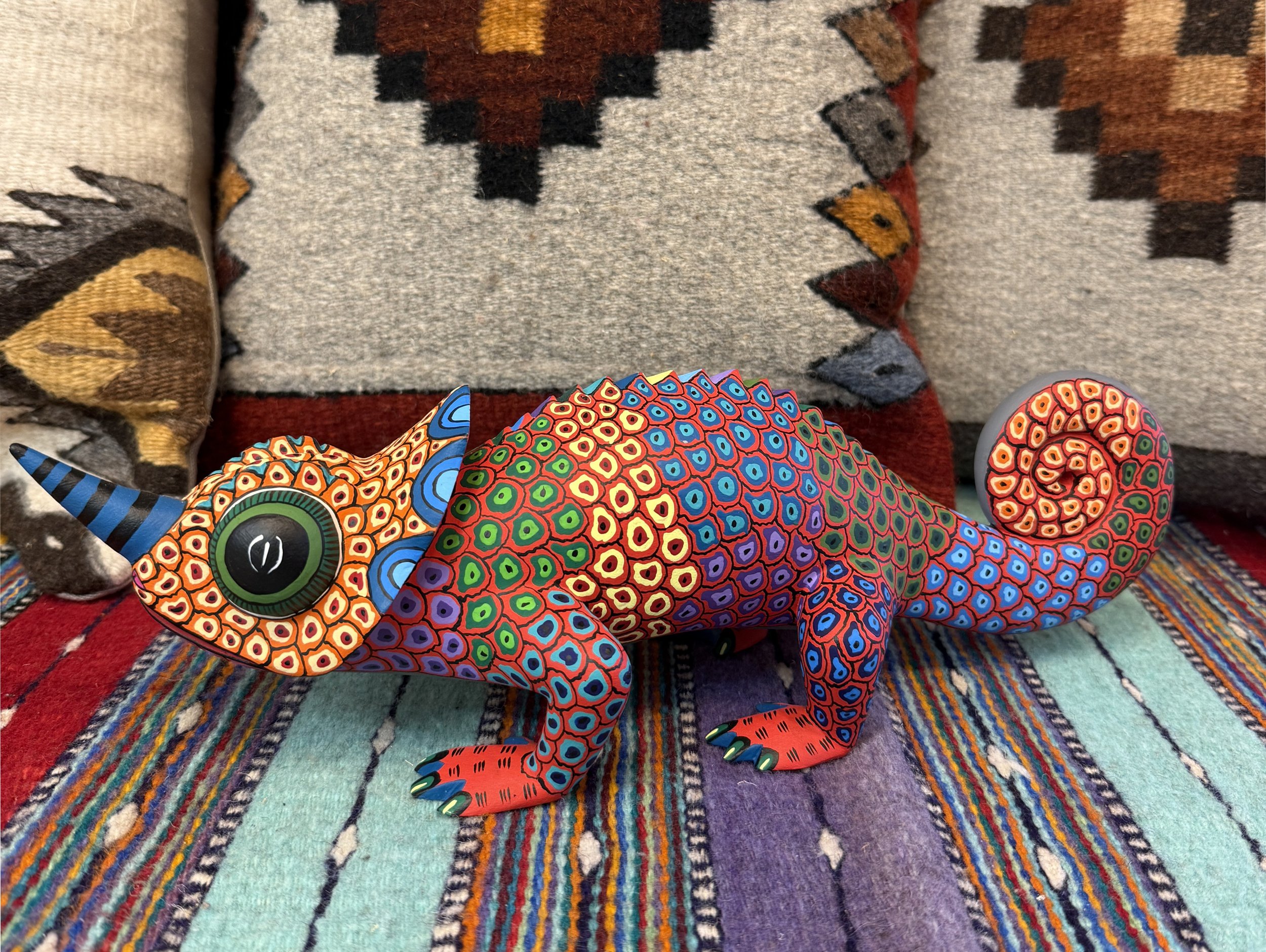 A colorful, patterned chameleon figurine with a spiral tail, large green eye, and a striped horn, resting on a multicolored woven fabric with a southwestern-style cushion in the background.