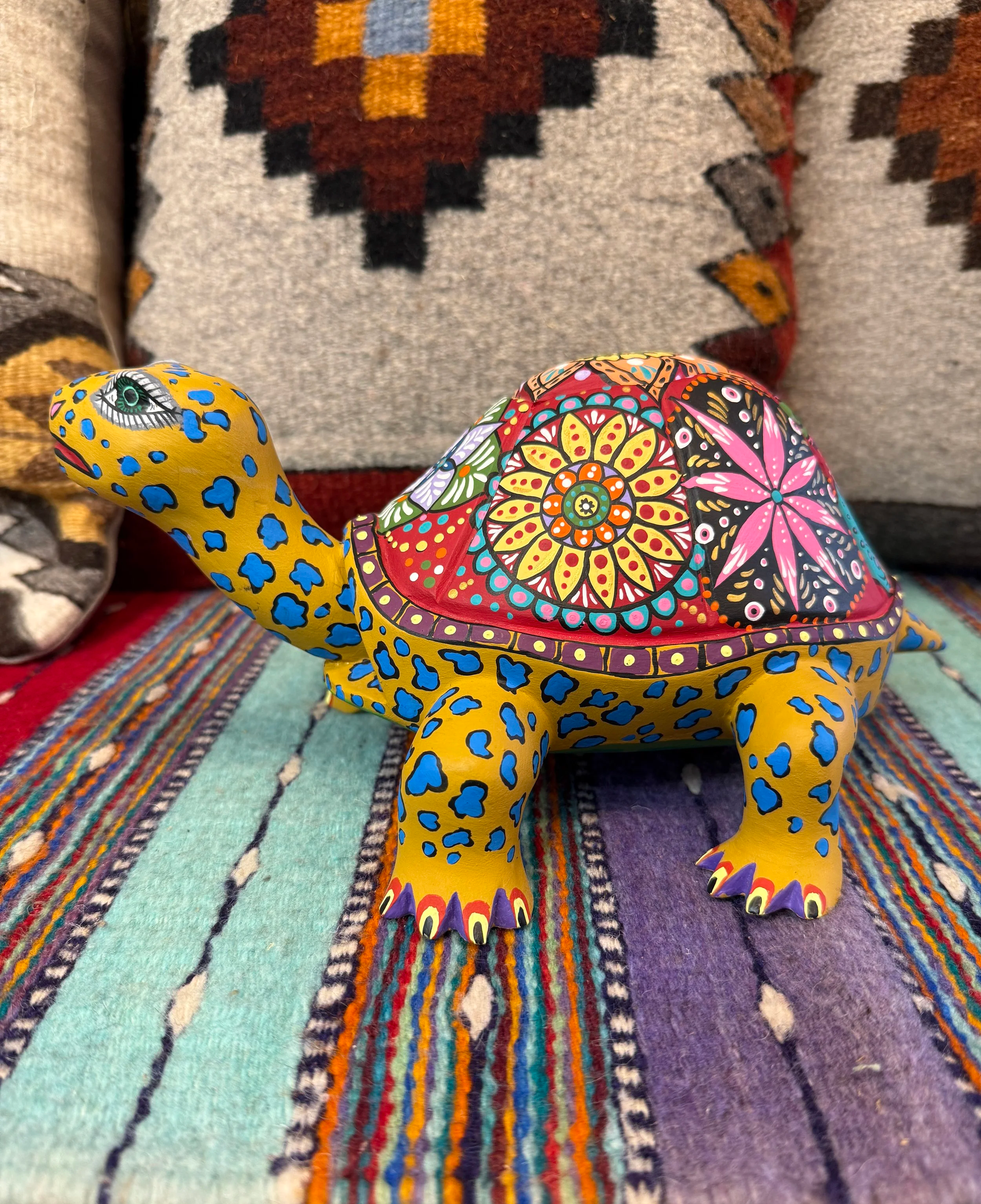 Colorful, decorated turtle figurine with a vibrant, patterned shell and yellow body with blue spots, placed on a colorful striped fabric with cushions in the background.