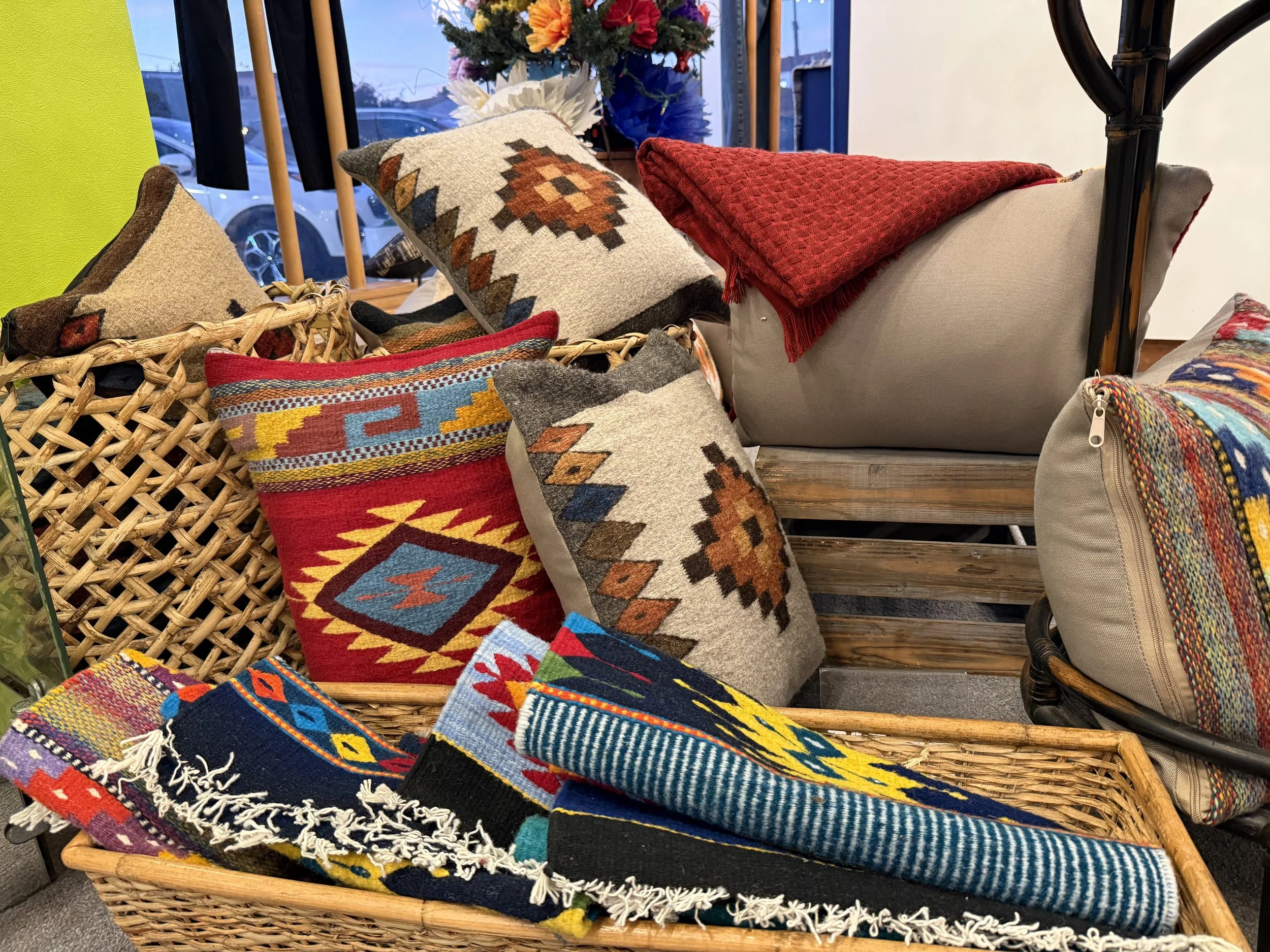 A display of colorful woven and decorative pillows, blankets, and textiles arranged in baskets and on a wooden bench inside a store.
