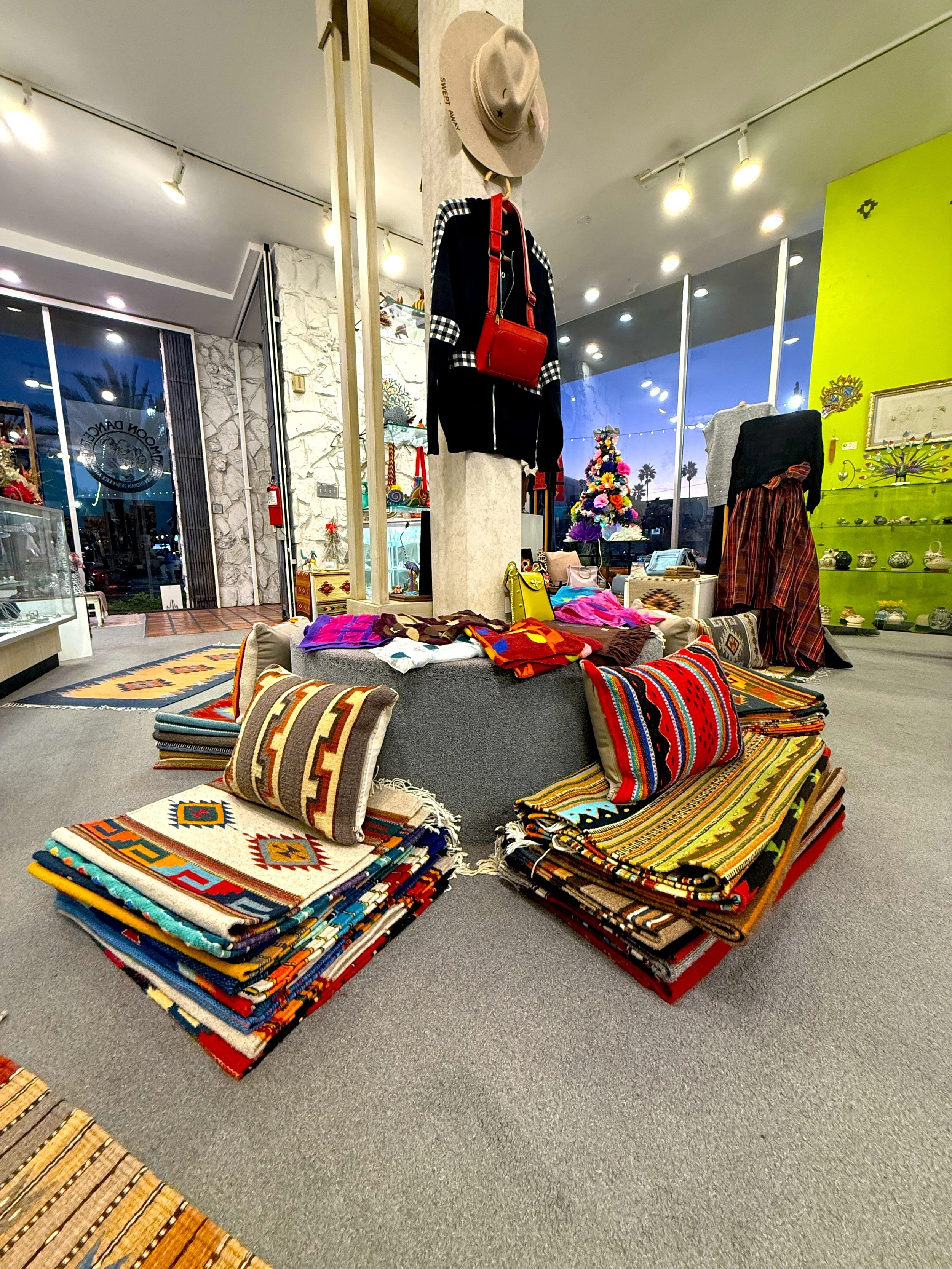 Display of colorful woven blankets, pillows, and clothing inside a store, including garments and accessories, large window showing Los Angeles palm trees.