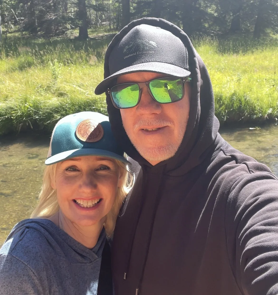 Smiling couple outdoors near a stream, taking a selfie on a sunny day. The woman has blonde hair and is wearing a teal hat and gray hoodie. The man has facial hair, sunglasses, a black hoodie, and a black cap.