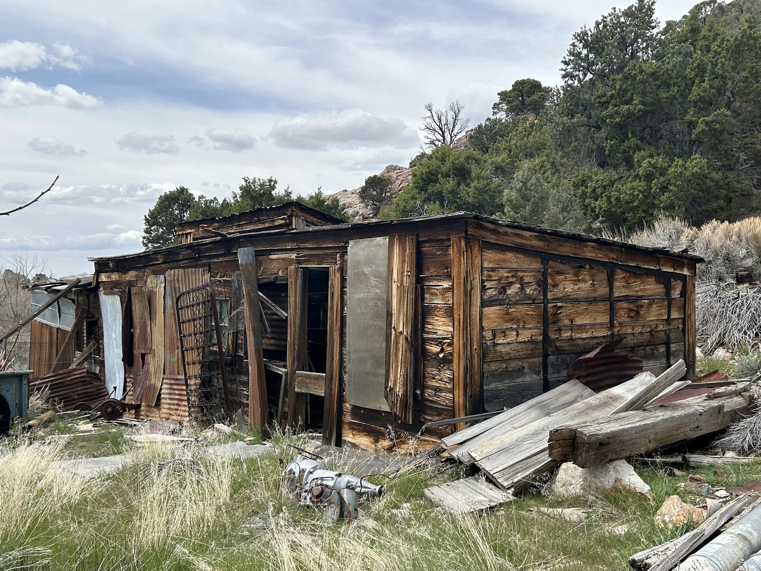 Osceola Nevada, Ghost town An old, dilapidated wooden shed with broken and missing panels, surrounded by scattered debris and overgrown grass, set against a hilly background with trees and cloudy sky.