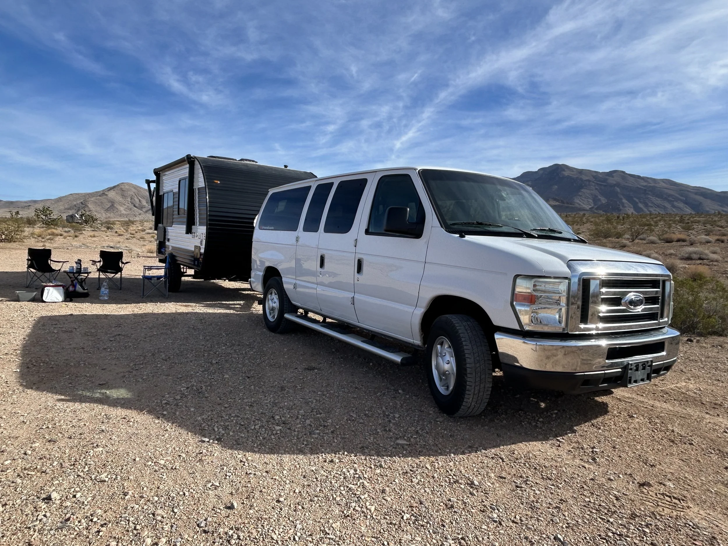 Van Life , Boondocking. White van Ford E350 and a travel trailer parked on a desert landscape with mountains in the background. There are two camping chairs, a small table, and camping supplies nearby under a clear blue sky.