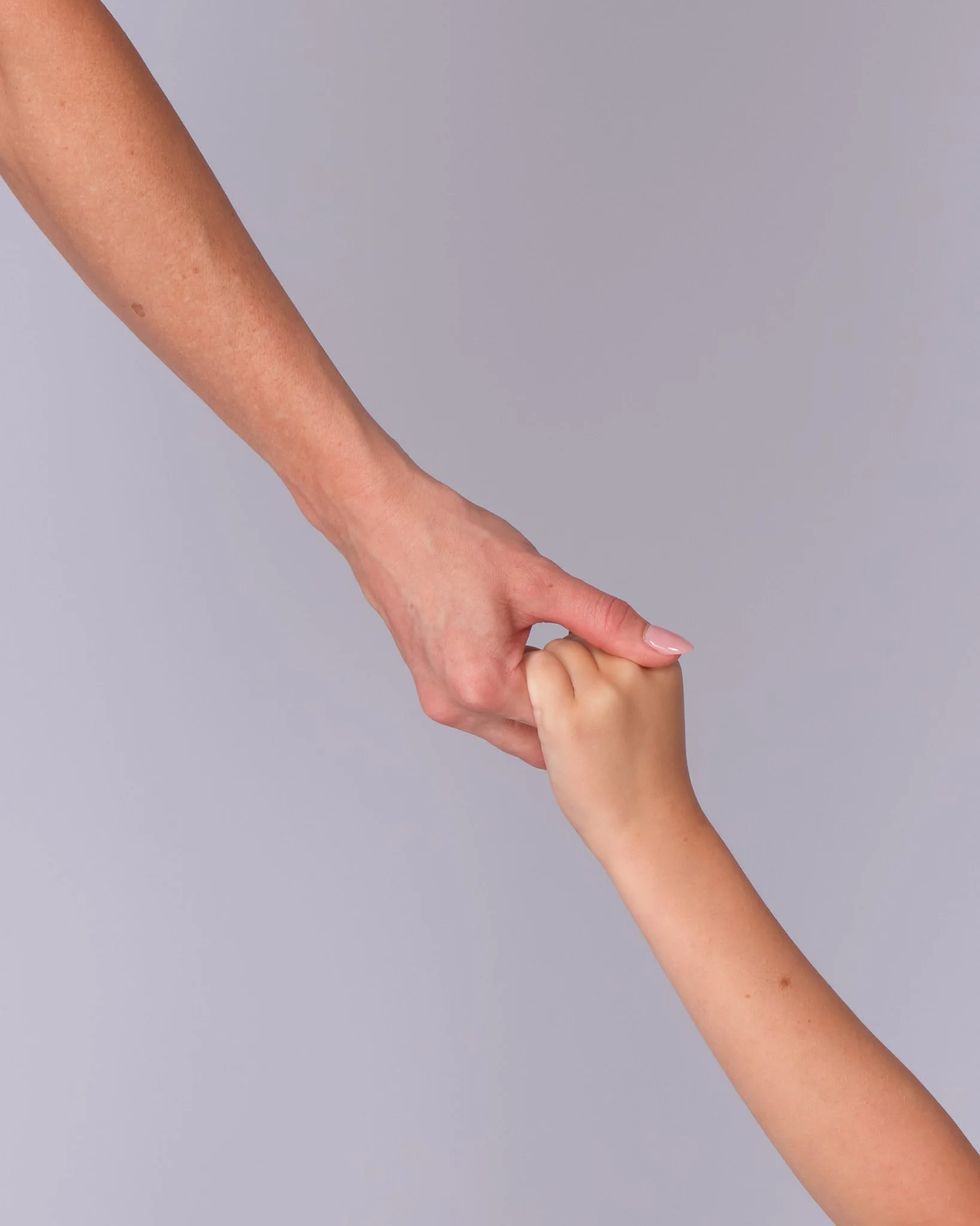 A person with a lighter skin tone holding a child's hand with a slightly darker skin tone against a plain light gray background.