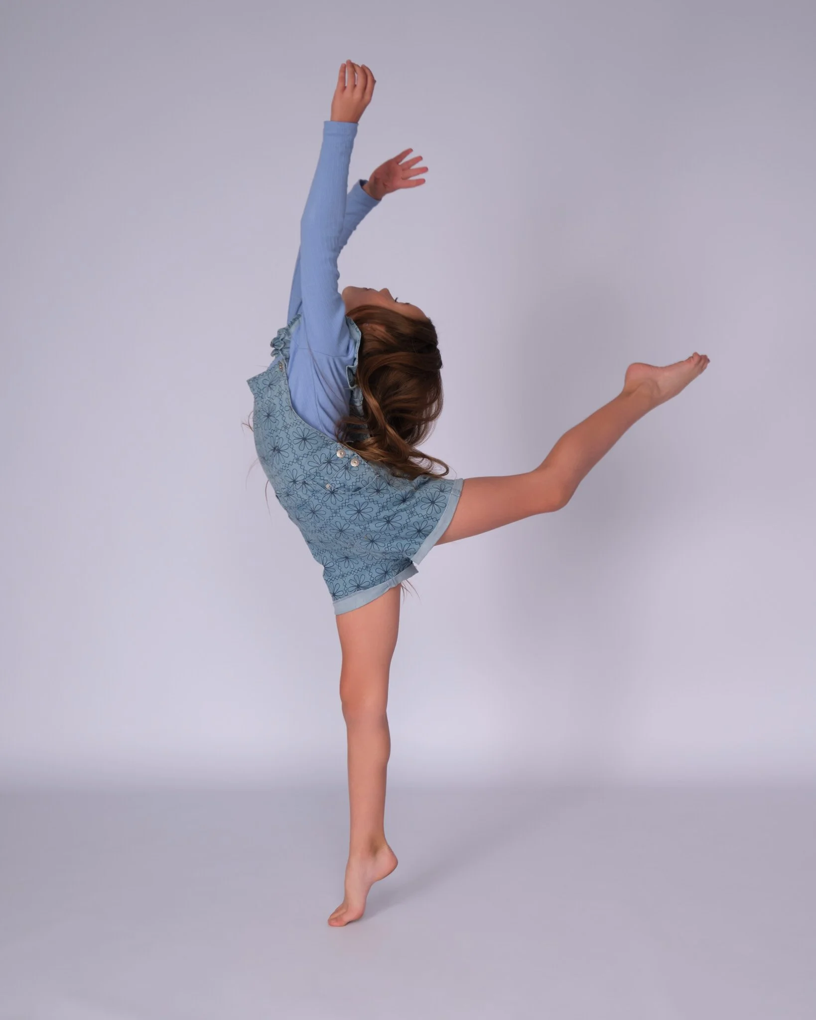 A young girl in a blue dress performing a handstand or gymnastic pose against a plain background.