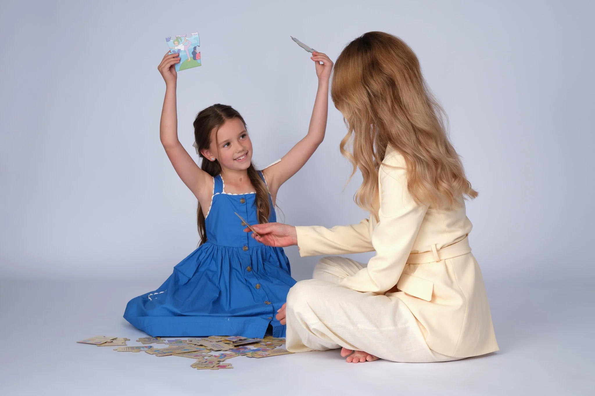 A young girl in a blue dress sits on the floor, smiling and holding up a puzzle piece. An adult woman in a light-colored outfit sits on the floor facing her, holding a piece of the puzzle. There are puzzle pieces scattered on the floor.