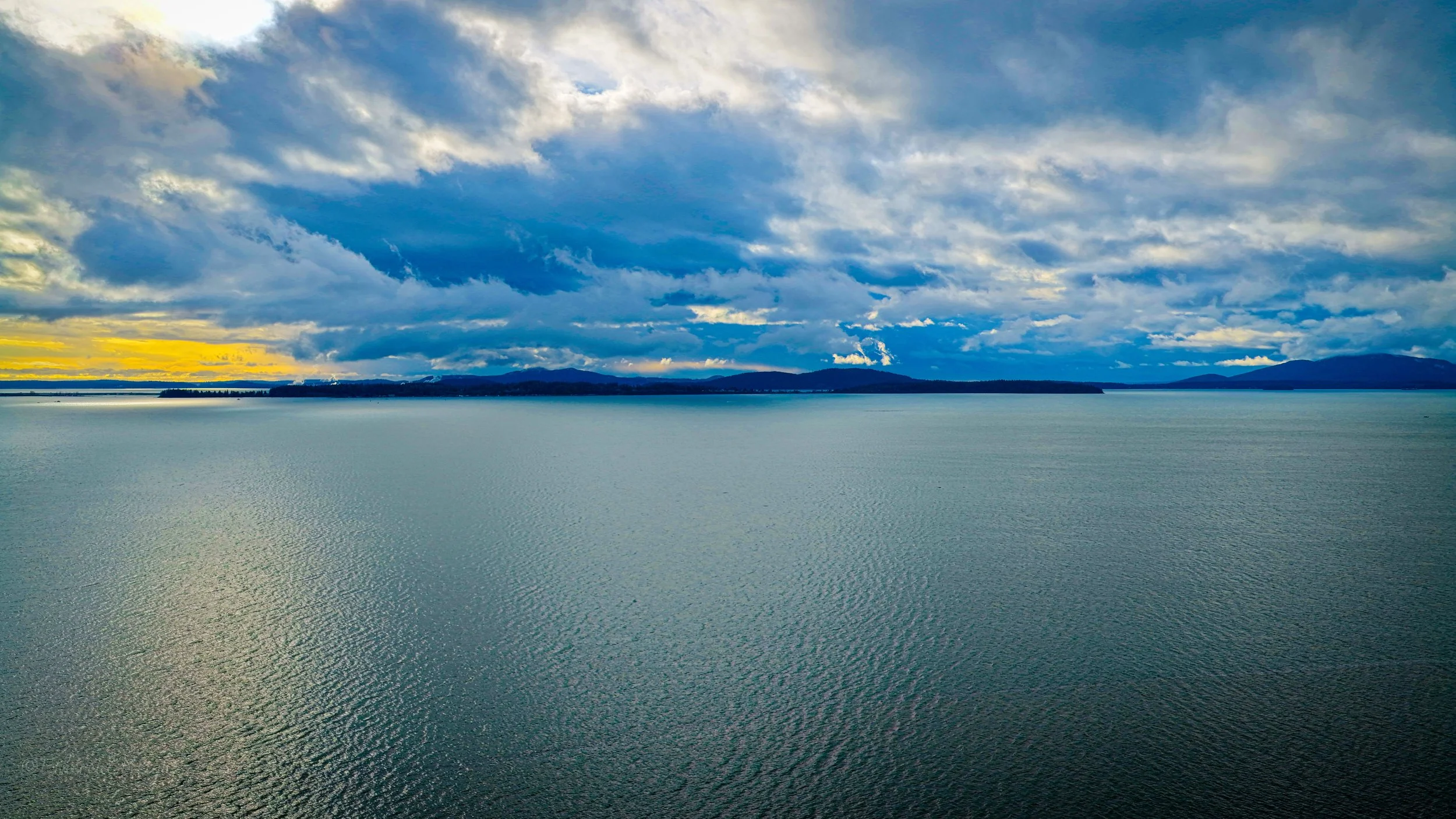 A vast body of water under a partly cloudy sky during sunset, with distant landforms and mountains on the horizon.