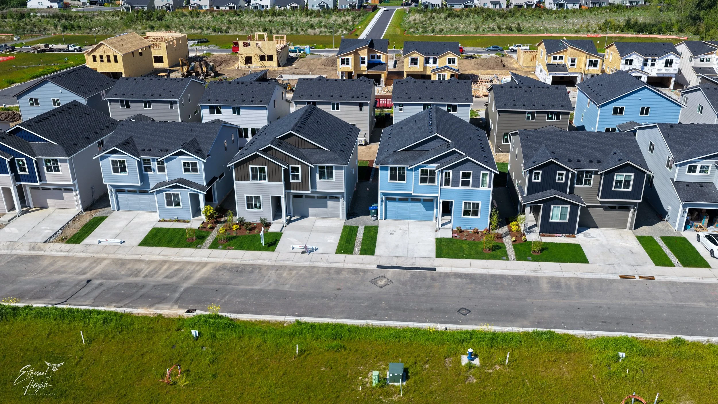 Aerial view of a suburban neighborhood with newly built blue, gray, and dark-colored houses in the foreground, and partially constructed houses with exposed wood framing in the background. The scene shows construction activity and parked vehicles.