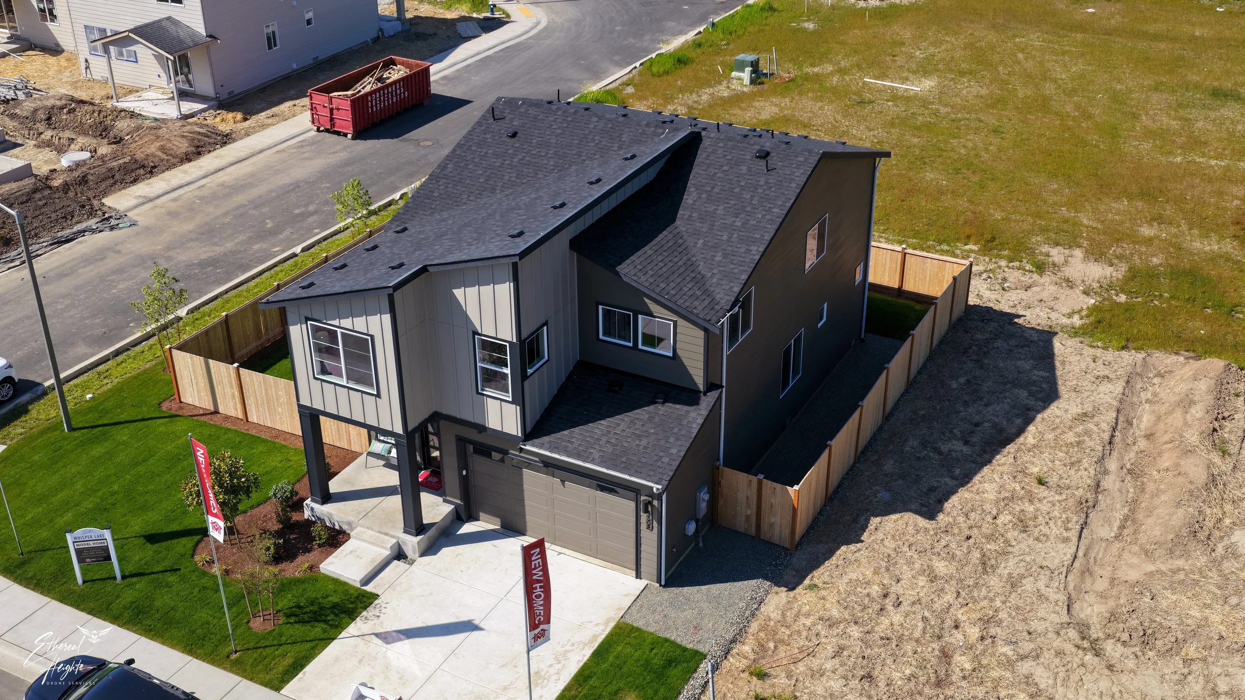A new house with a gray exterior, black roofing, and a fenced backyard. The house has multiple windows, a front porch, and a driveway. There are signs indicating it is a model home, with a small green lawn and a tree.