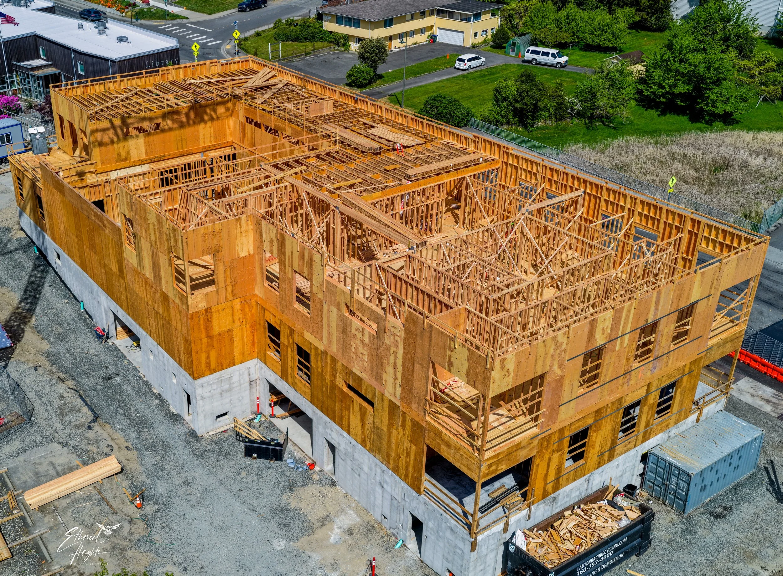 Aerial view of a multi-story building under construction with exposed wooden framing. Surrounding area includes parking lot, green lawn, trees, and nearby residential buildings.
