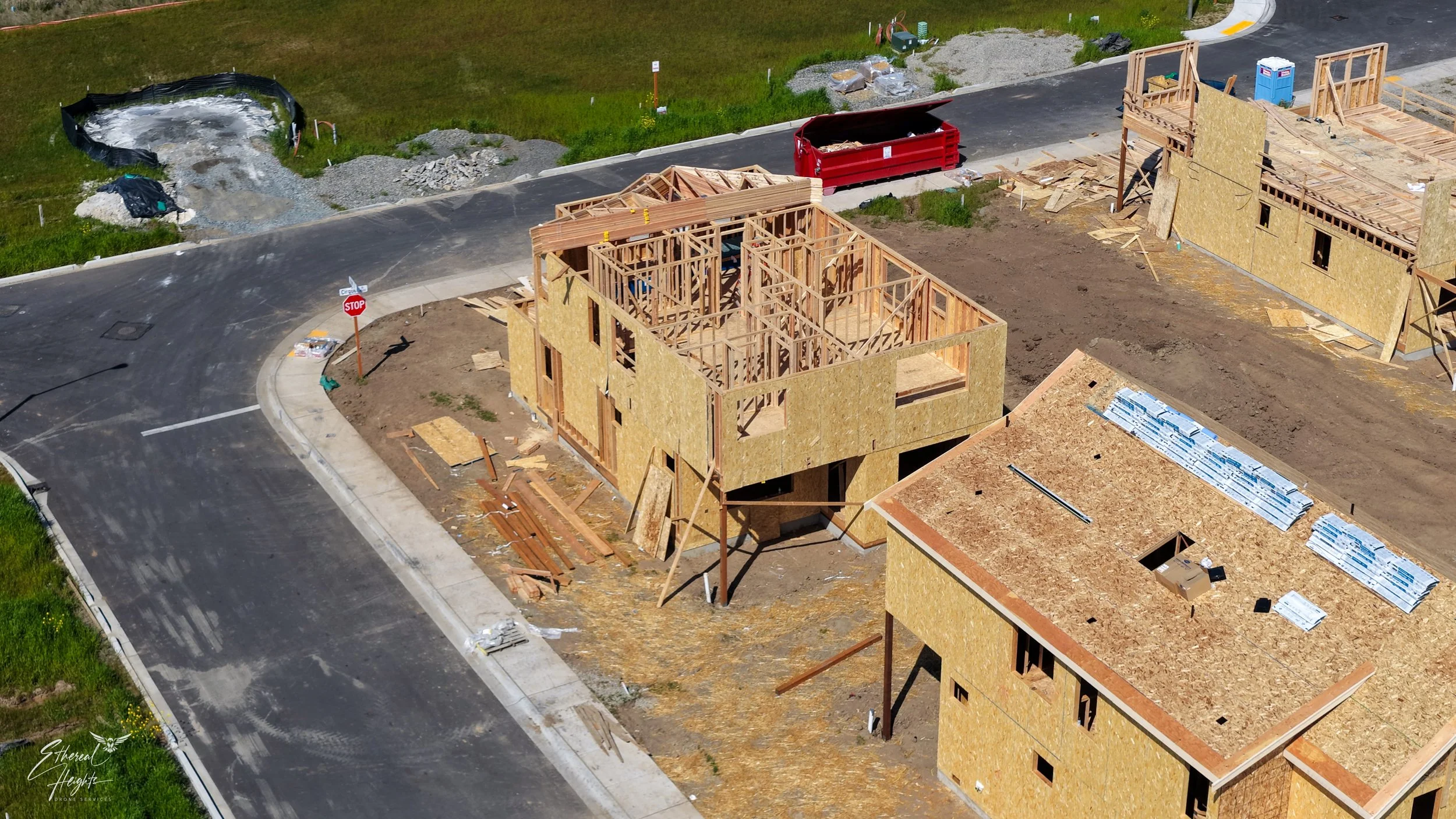 Construction site with wooden framing for new houses, surrounded by roads and grassy areas, some construction materials and equipment present.