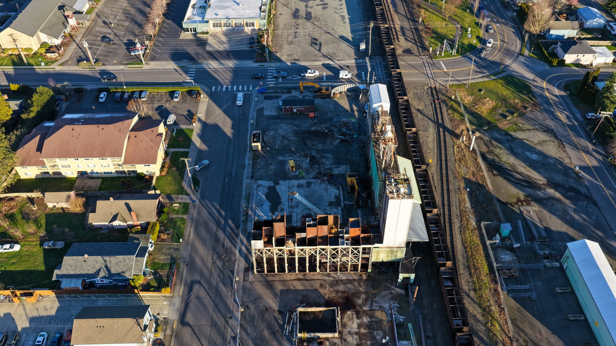 Aerial view of a construction site with a partially built structure, surrounded by roads, parked cars, residential houses, and a railway track.