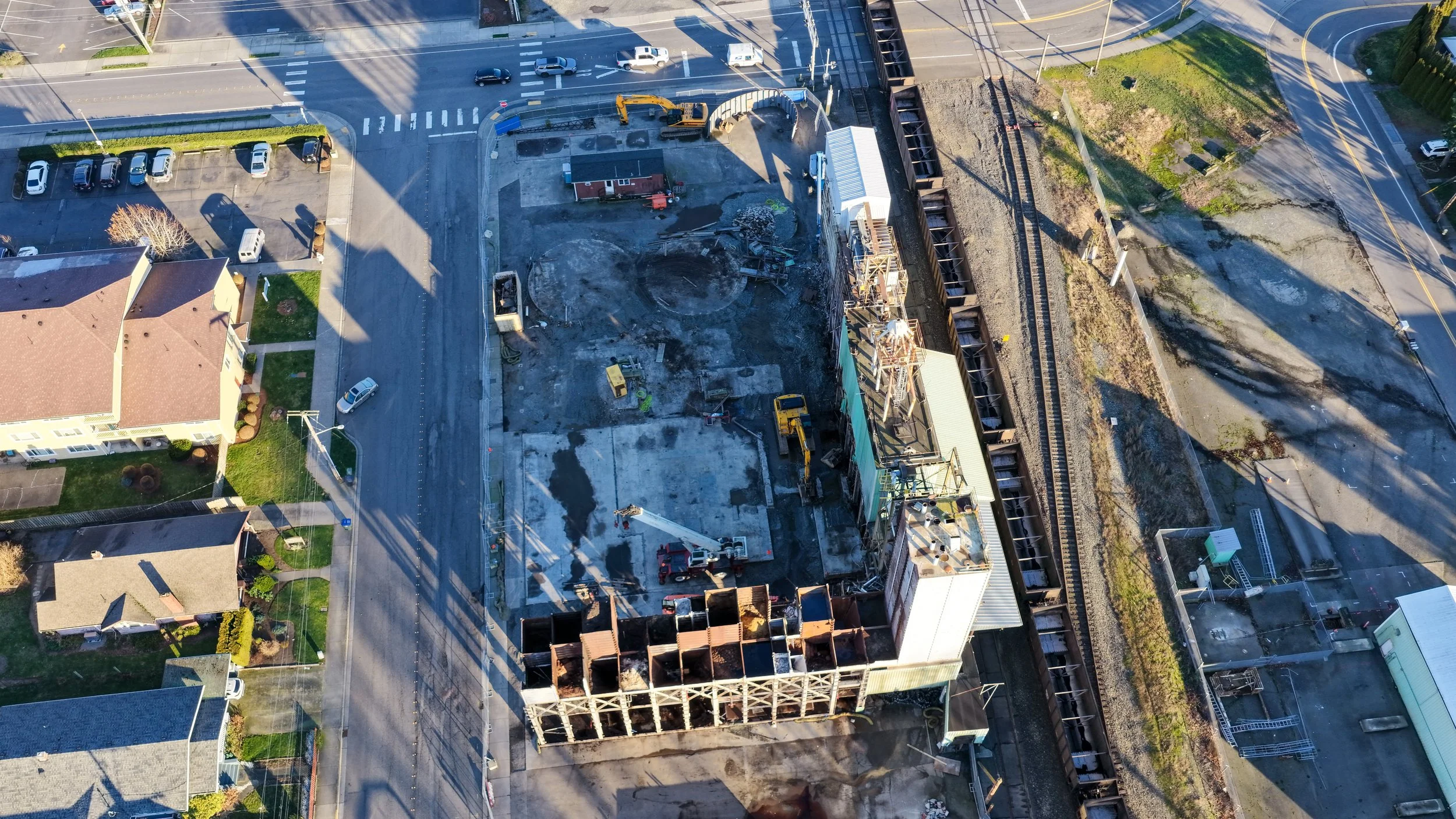 Aerial view of a construction site with machinery, partially built structures, and surrounding streets with parked cars and nearby buildings.